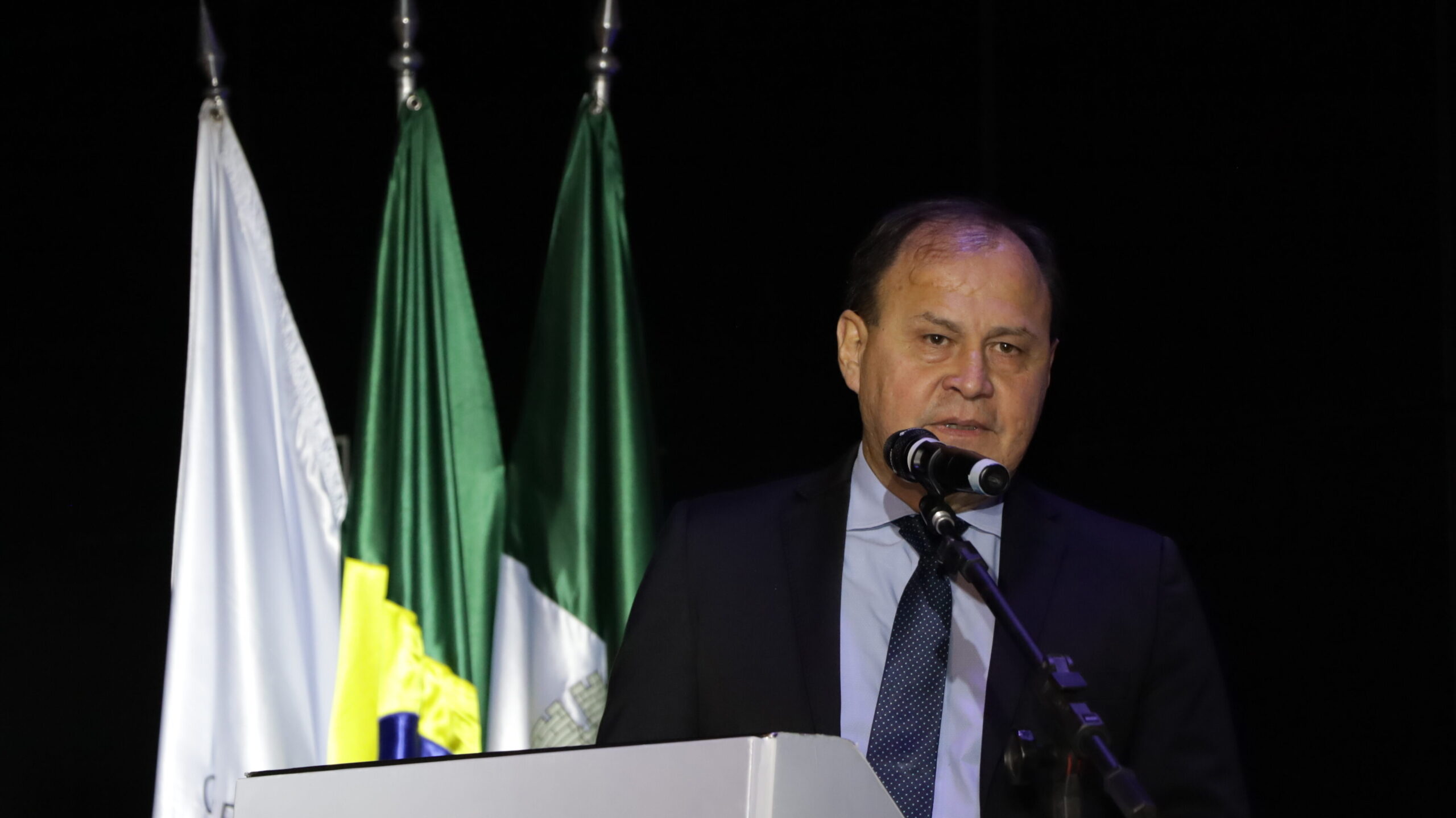 Um homem de terno preto e gravata azul-escura está em um pódio, falando em um microfone. Ao fundo, três bandeiras estão visíveis.
