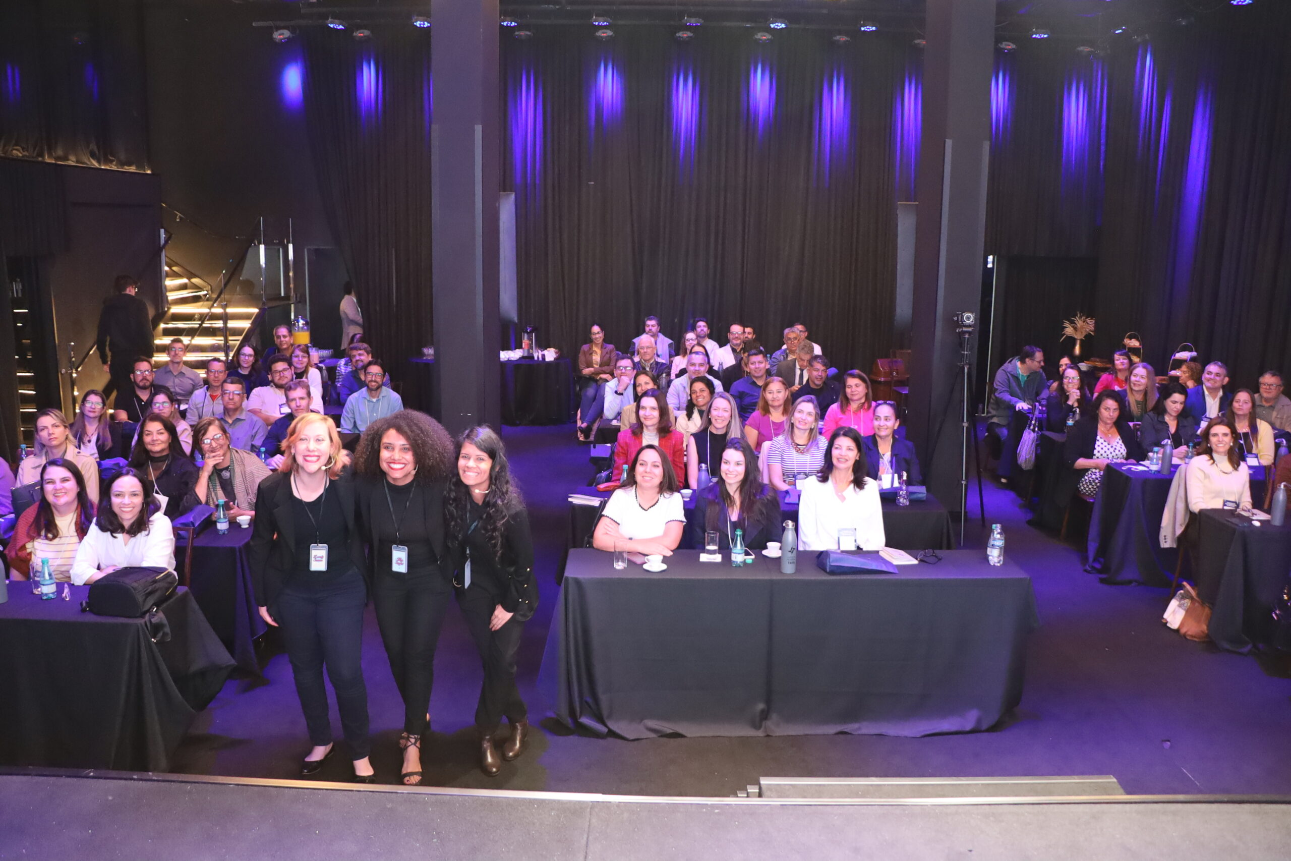 Uma foto de uma plateia em um evento, com fileiras de pessoas sentadas em mesas em um ambiente interno com iluminação baixa, olhando para a frente.