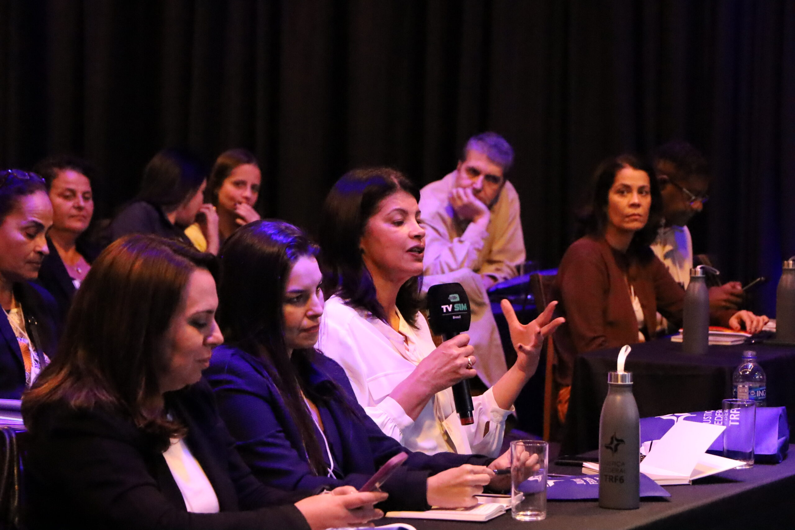 Uma foto de um grupo de pessoas sentadas em uma mesa, vestidas com trajes sociais, interagindo e participando de um painel ou discussão.