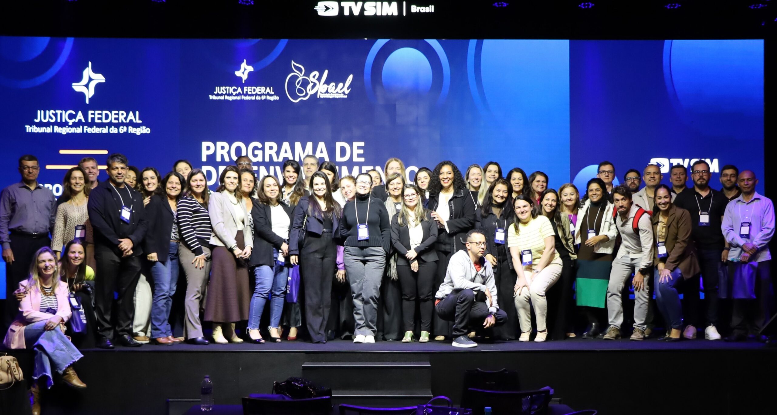 Um grande grupo de pessoas, vestidas de forma casual e semi-formal, está posando no palco à frente de uma tela iluminada. A tela exibe logotipos e o título "PROGRAMA DE DESENVOLVIMENTO DE GESTORES".