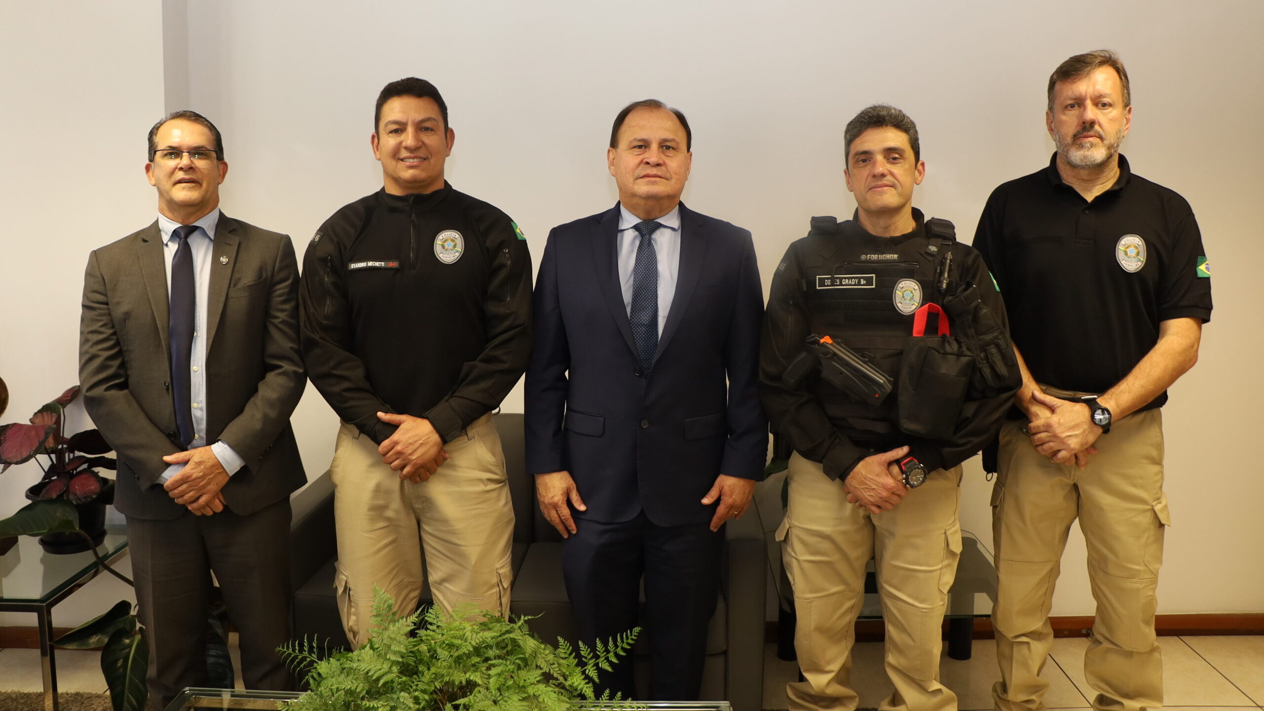 Cinco homens posam lado a lado em um ambiente interno. Dois homens vestem terno e gravata, e os outros três vestem uniforme preto e calças bege. Todos olham diretamente para a câmera.