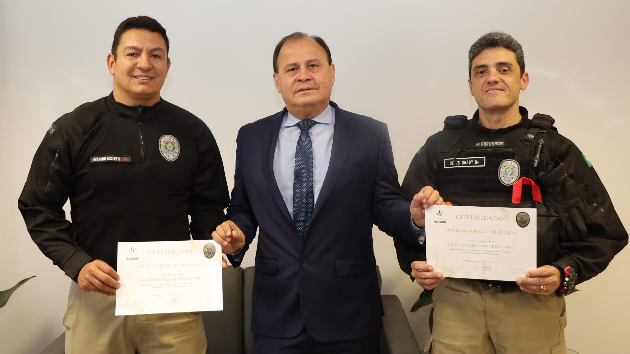 A imagem mostra três homens posando lado a lado em um ambiente interno. O homem da esquerda e o da direita vestem uniformes de cor preta e seguram um certificado individualmente, ambos exibindo o brasão da Polícia Judicial. O homem no centro veste um terno azul-marinho com gravata azul. Os três indivíduos olham diretamente para a câmera, com expressões sorridentes.