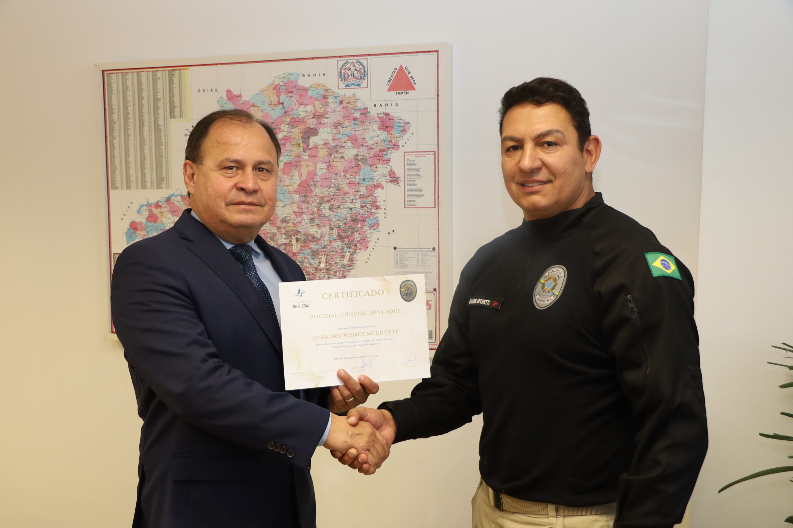 Um homem de terno azul e um homem de uniforme preto se cumprimentam com um aperto de mãos, enquanto o homem de terno entrega um certificado com a mão esquerda. Ao fundo, pode-se ver um mapa fixado na parede.