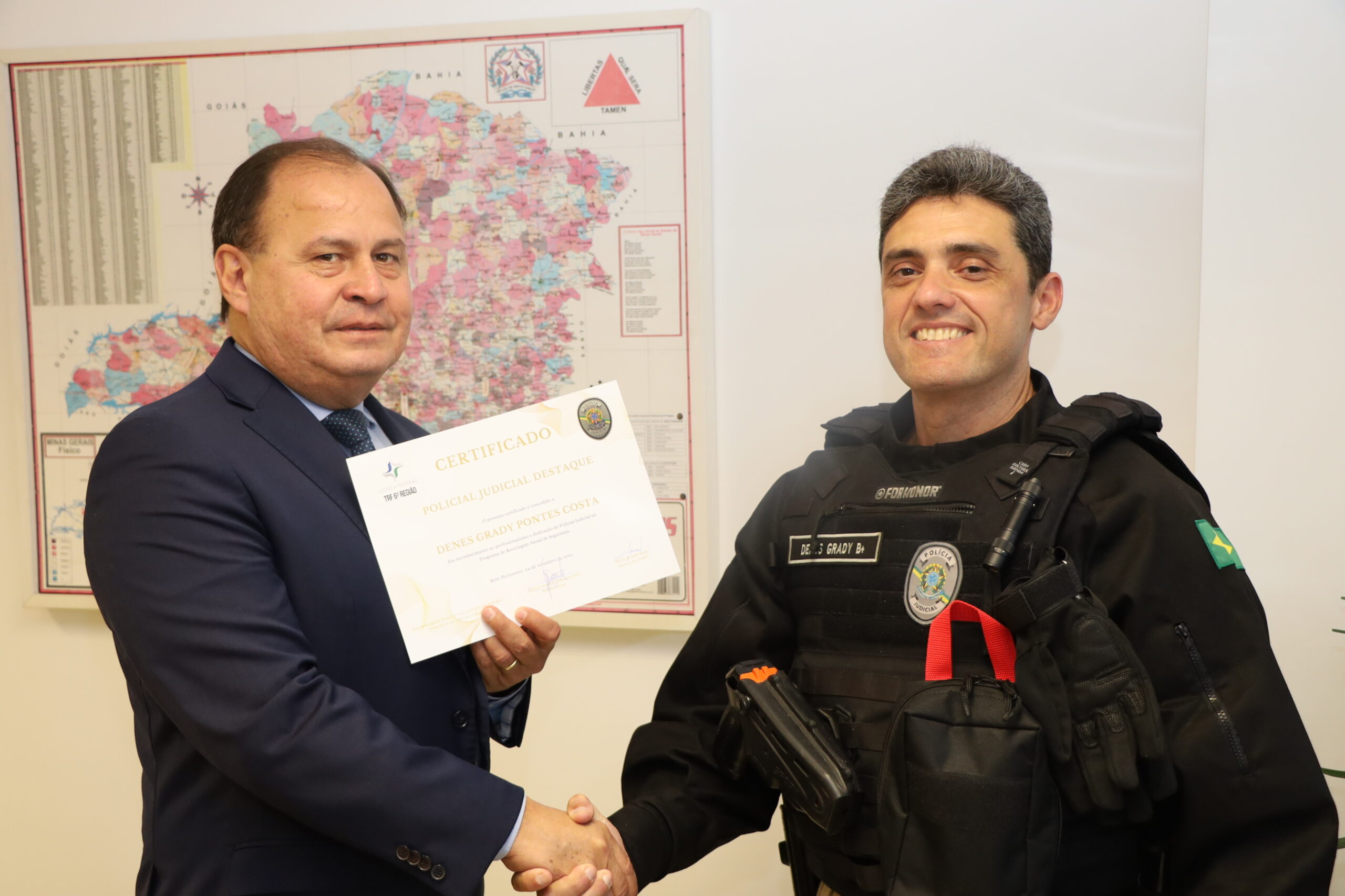 Um homem de terno azul e um homem de uniforme preto se cumprimentam com um aperto de mãos, enquanto o homem de terno entrega um certificado. Eles estão de pé, com um mapa e uma parede clara ao fundo.