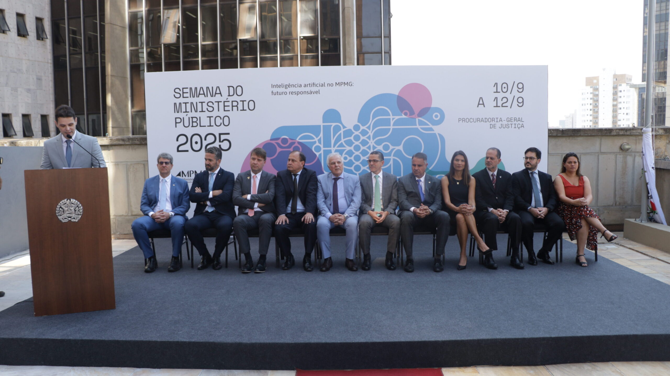 Cena de evento externo, com um homem em pé discursando em um púlpito com o brasão da República, e um grupo de onze pessoas sentadas (nove homens e duas mulheres) na primeira fila. Todos vestem trajes formais ou sociais.