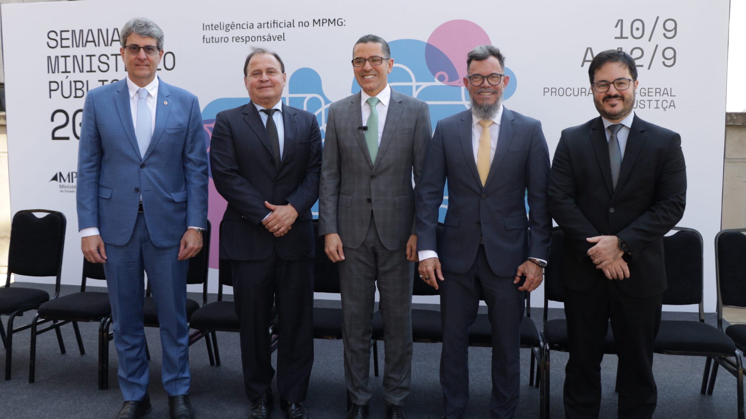Foto de grupo de cinco homens posando lado a lado em um evento externo, todos vestindo trajes formais (ternos e gravatas). Eles estão em pé na frente de cadeiras vazias, com um painel de fundo que contém o tema do evento.