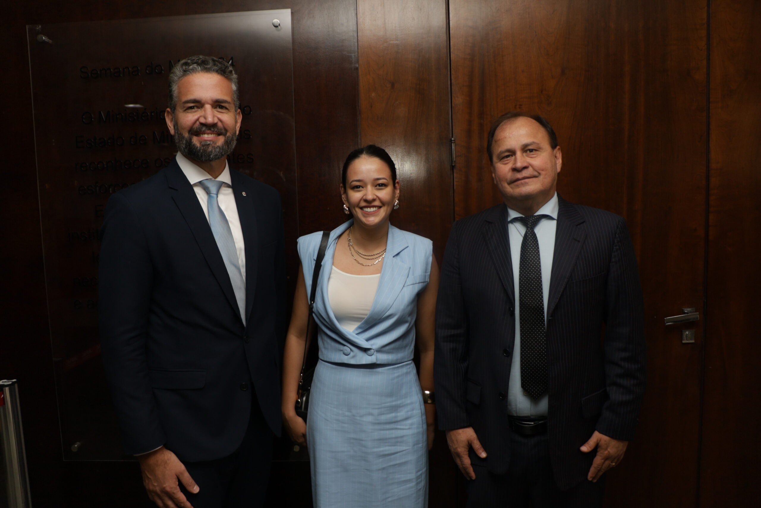 Grupo de três pessoas (dois homens e uma mulher) em ambiente interno, posando em frente a uma porta de madeira. Os homens vestem trajes formais (ternos) e a mulher veste um conjunto social.
