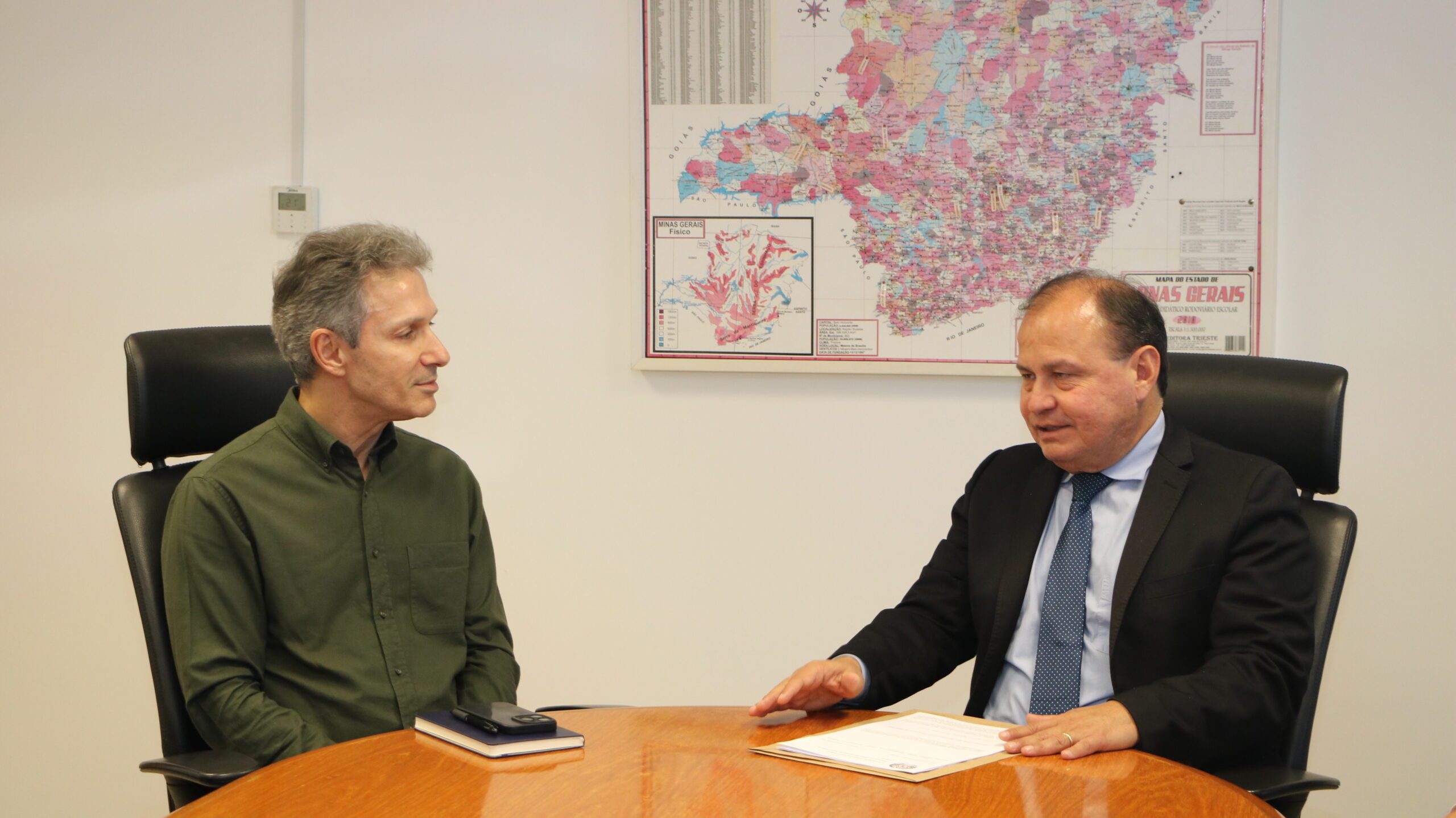 A imagem mostra duas pessoas sentadas em uma mesa redonda de madeira. O homem à esquerda, de cabelo grisalho, usa uma camisa de cor verde-oliva. O homem à direita veste um terno escuro e uma gravata azul-clara. Ambos estão voltados um para o outro, conversando. O homem de terno gesticula com a mão direita. Ao fundo, em uma parede branca, há um grande mapa colorido de Minas Gerais.