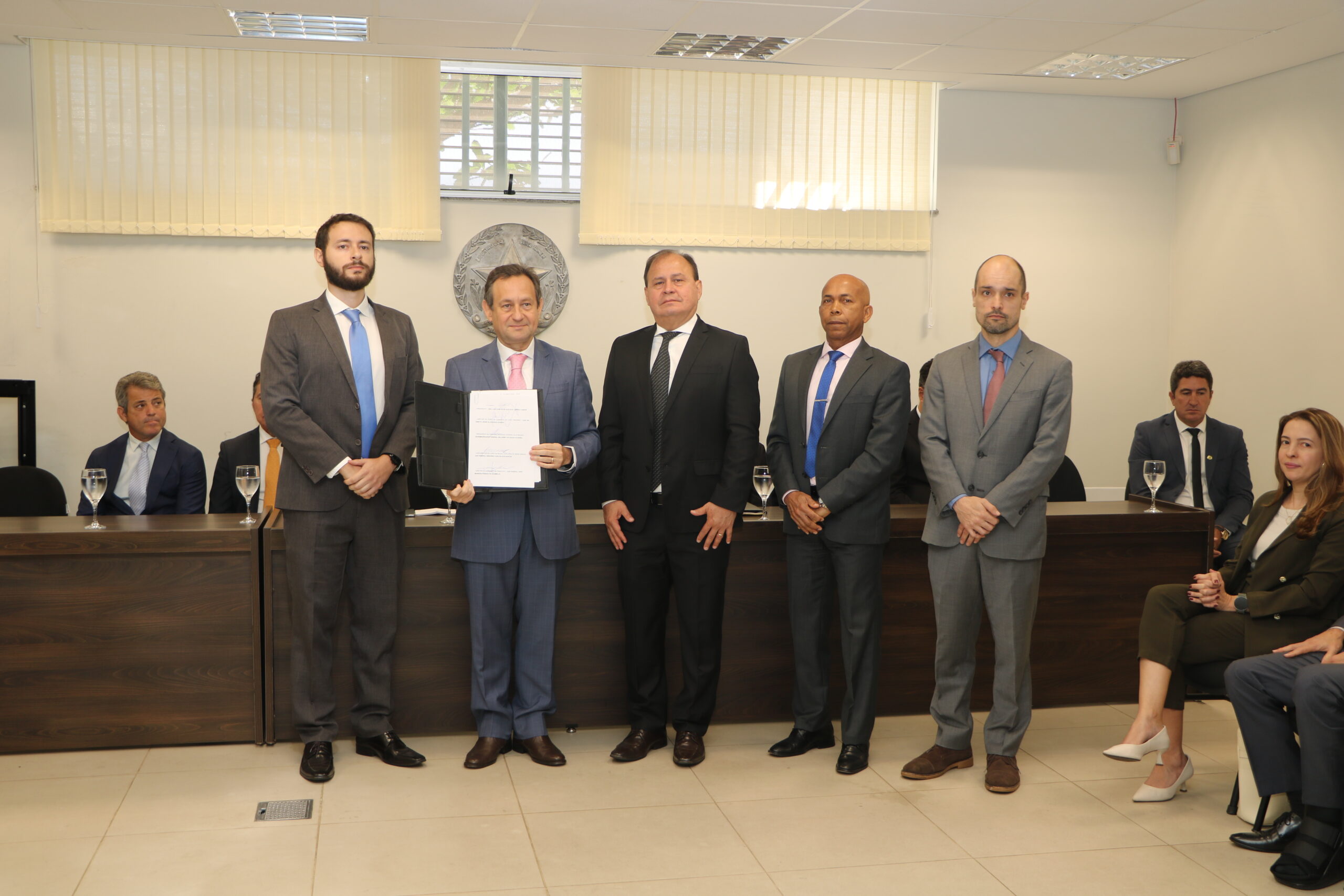 Cena de um evento formal com um grupo central de cinco homens em pé, todos em vestes formais (ternos), com um dos homens segurando um documento. Outras pessoas, sentadas ou paradas, estão visíveis no fundo e nas laterais. O ambiente é uma sala de reuniões com janelas ao fundo.