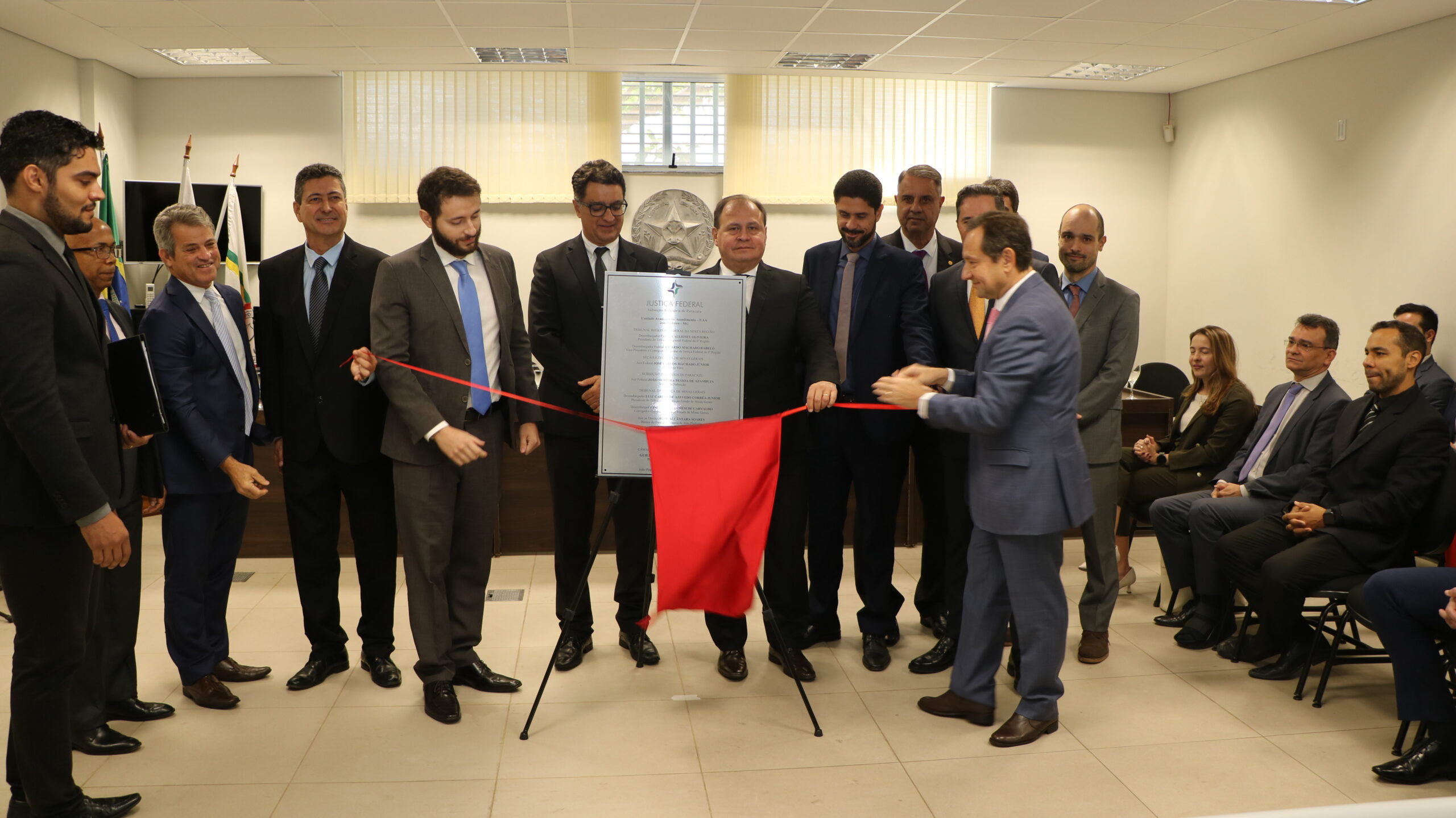 Cerimônia de inauguração com descerramento de placa e corte de fita, mostrando um grupo de homens em trajes formais (ternos) ao redor de uma placa metálica coberta por um tecido. Dois homens nas laterais da placa seguram a fita, e há outras pessoas sentadas na lateral direita.