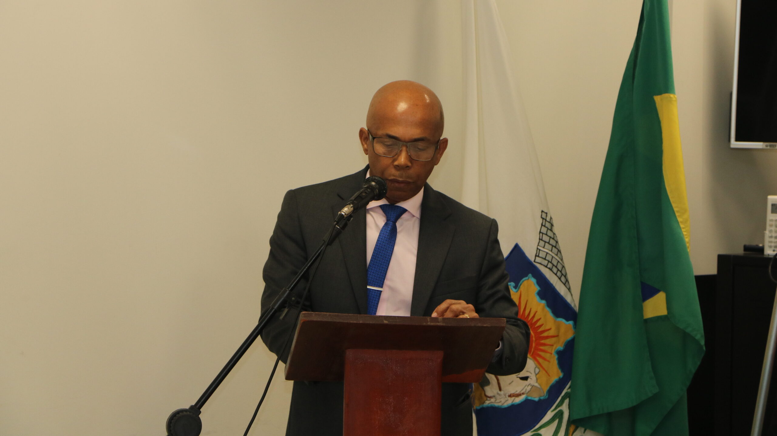 Homem careca, usando óculos e vestes formais (terno e gravata), inclinado sobre um púlpito de madeira enquanto fala em um microfone. Ele está em um ambiente interno com fundo claro, com bandeiras visíveis atrás.
