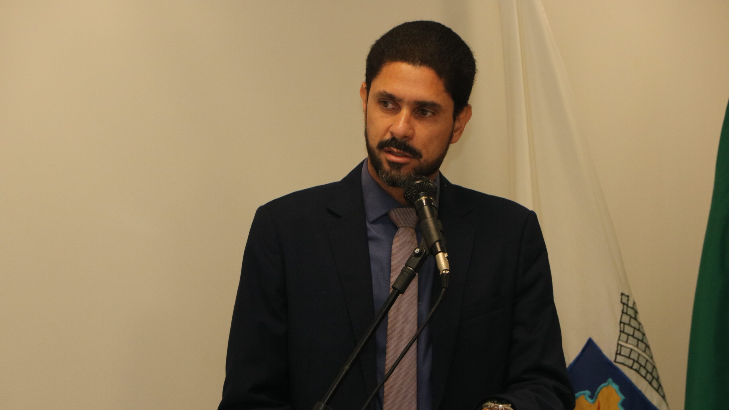Retrato de um homem com barba, em vestes formais (terno e gravata), falando em um microfone que está em primeiro plano. Ele está em um ambiente interno com fundo claro.