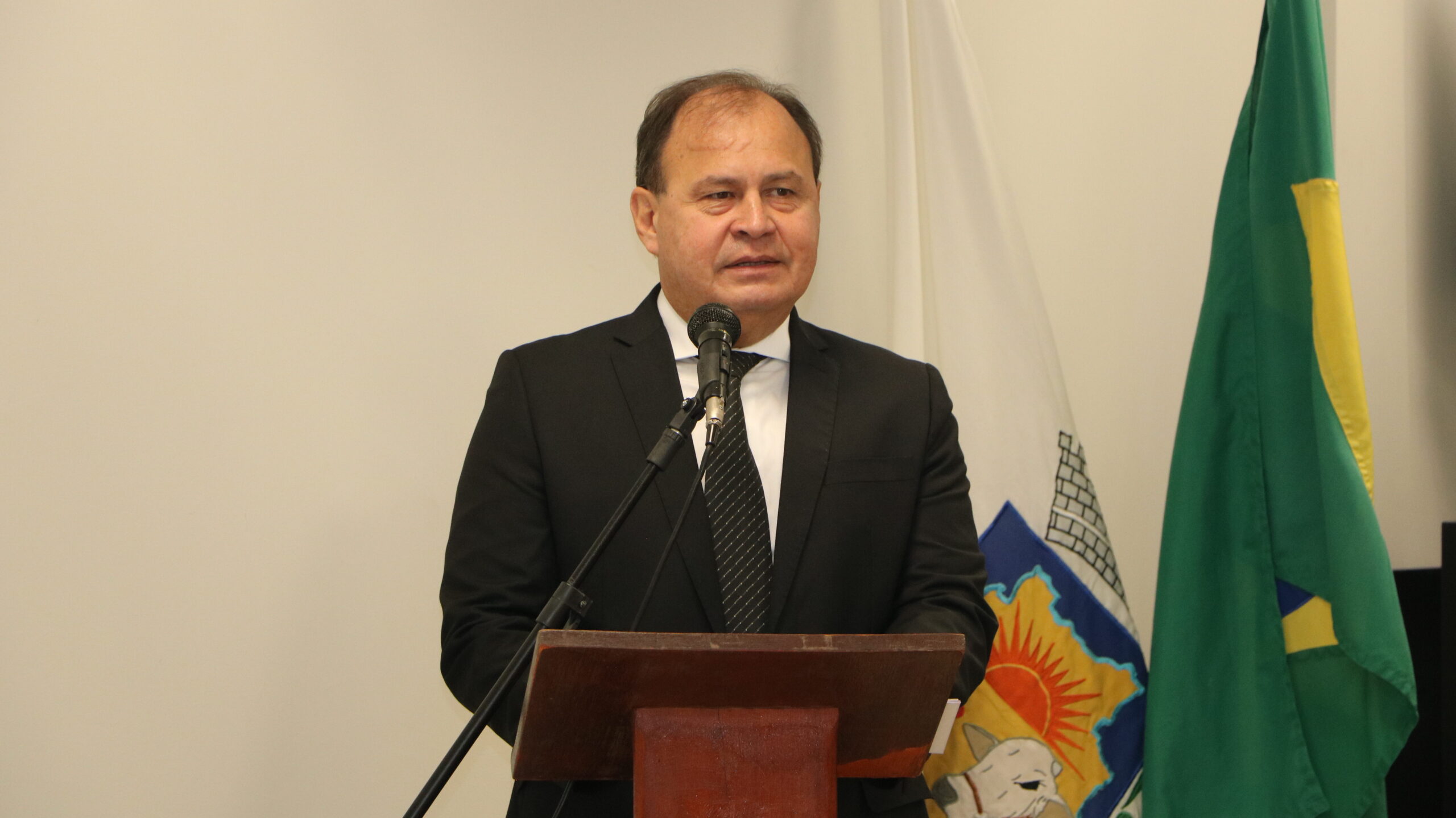 Retrato de um homem em discurso, usando vestes formais (terno e gravata), posicionado atrás de um púlpito de madeira com microfone. Ao fundo, duas bandeiras hasteadas estão parcialmente visíveis.