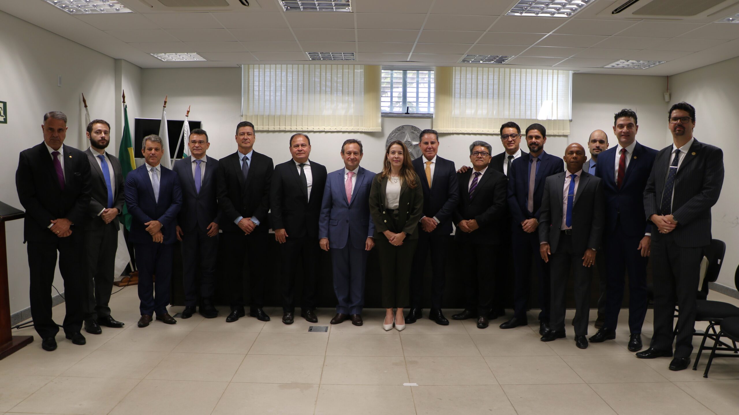 Foto de grupo de dezesseis pessoas (quinze homens e uma mulher) em um ambiente de sala de reuniões/sessões. Quase todos os homens vestem trajes formais (ternos) e a mulher veste calça e blazer. Ao fundo, há bandeiras e uma janela coberta por persianas.