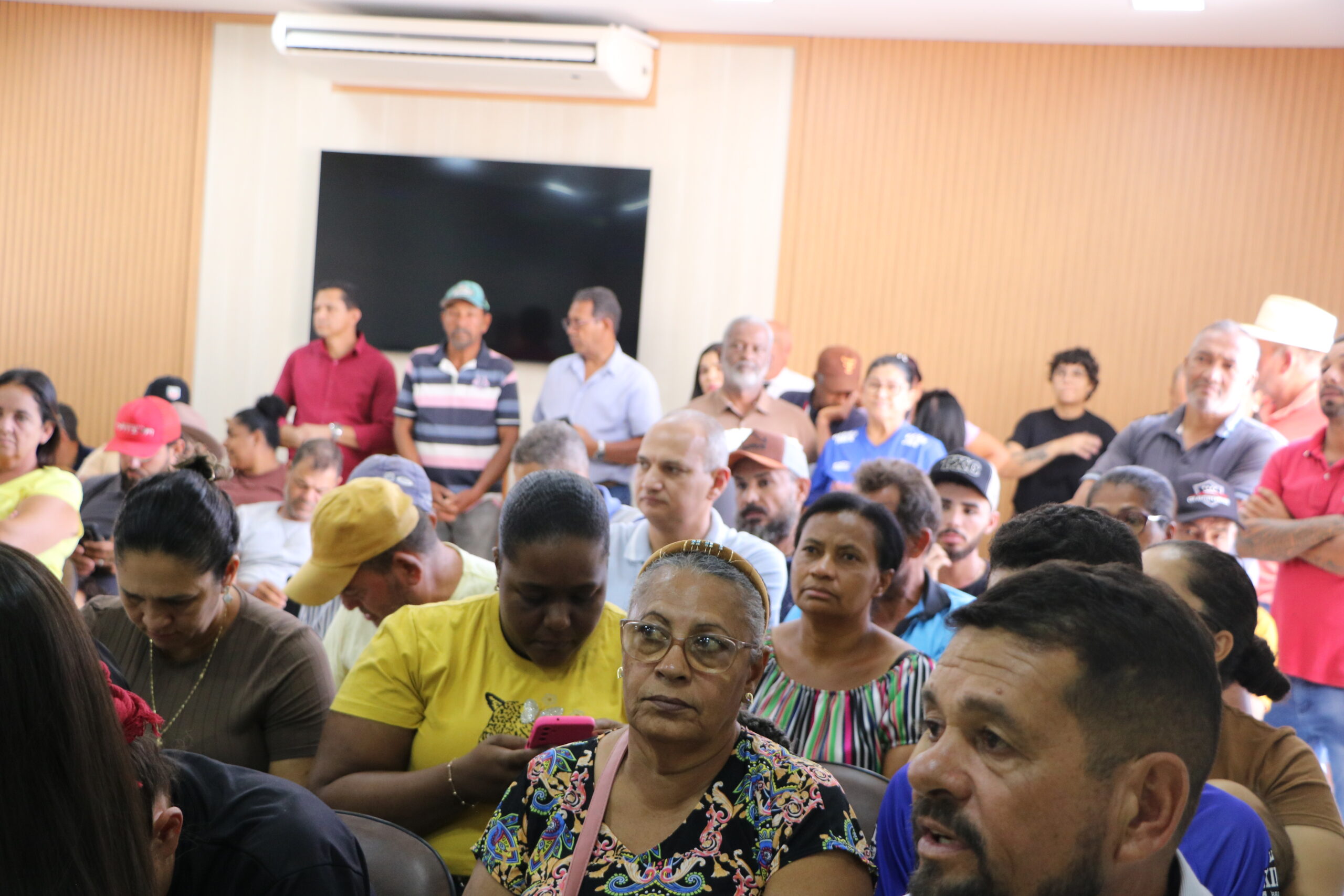 Um grande grupo de pessoas de diversas idades e etnias está reunido em um ambiente interno. As pessoas vestem trajes casuais e estão atentas, olhando para a frente. O fundo mostra uma parede revestida de madeira clara e uma tela escura.