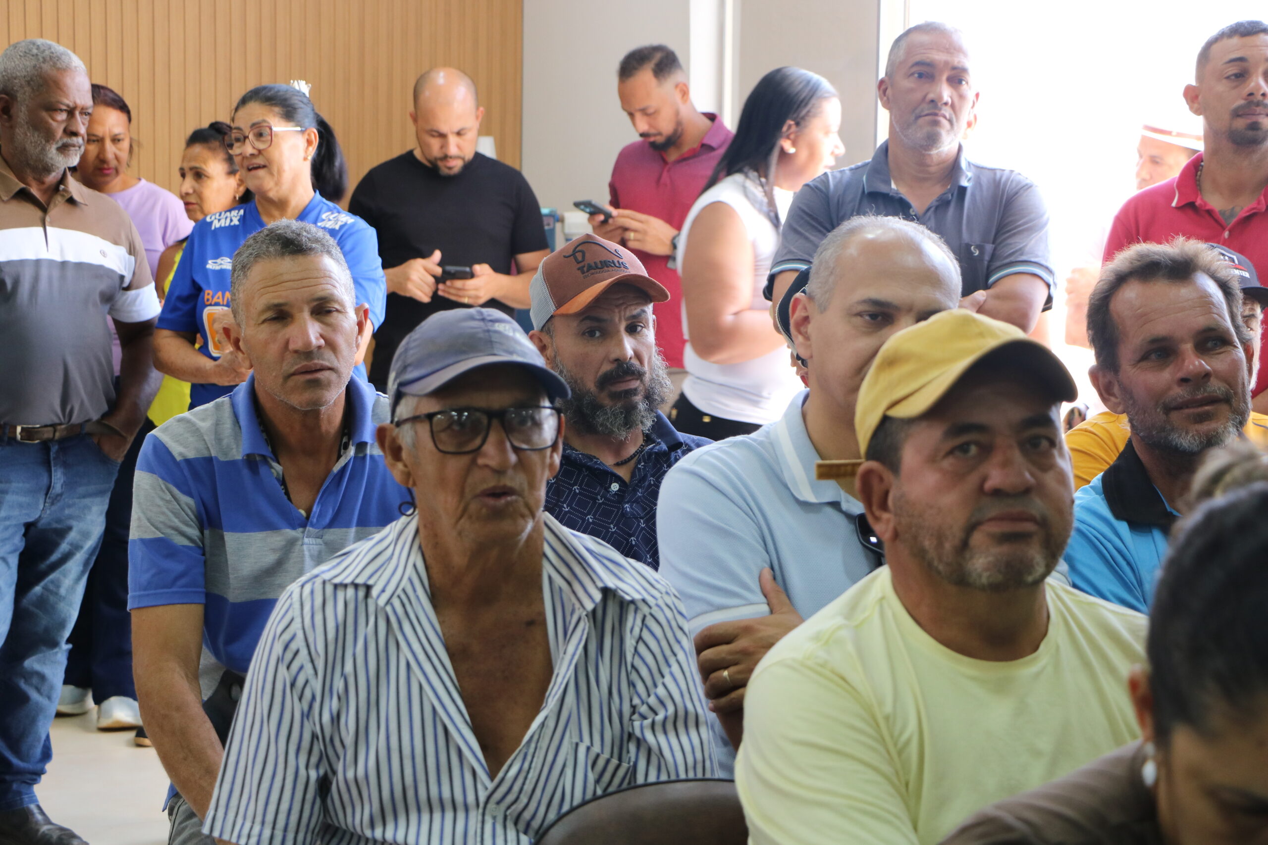 Um close-up de um grupo de pessoas, principalmente homens, em trajes casuais, sentadas ou em pé. Elas estão atentas, com expressões sérias, observando uma atividade fora do quadro.