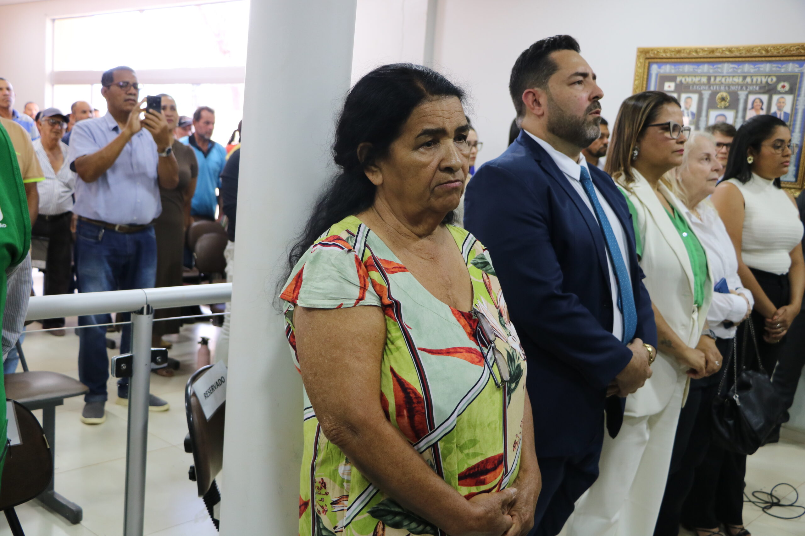 Um grupo de pessoas está reunido em um ambiente interno. No primeiro plano, uma mulher em trajes sociais coloridos está em destaque, ao lado de homens e mulheres em trajes formais e semi-formais. No fundo, outras pessoas, algumas com câmeras, estão observando a atividade.