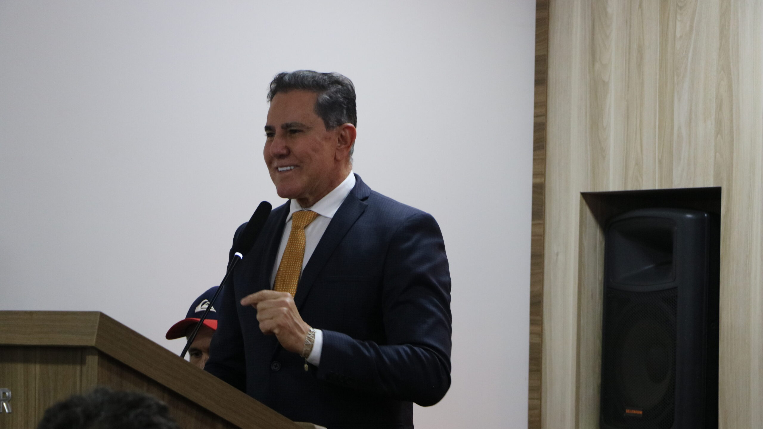 Um close-up de um homem sorrindo, vestido com traje formal, enquanto fala ao microfone em um púlpito de madeira. Ele está gesticulando com a mão direita. O fundo é simples, com uma parede branca e um painel de madeira à direita, onde um alto-falante escuro está embutido.