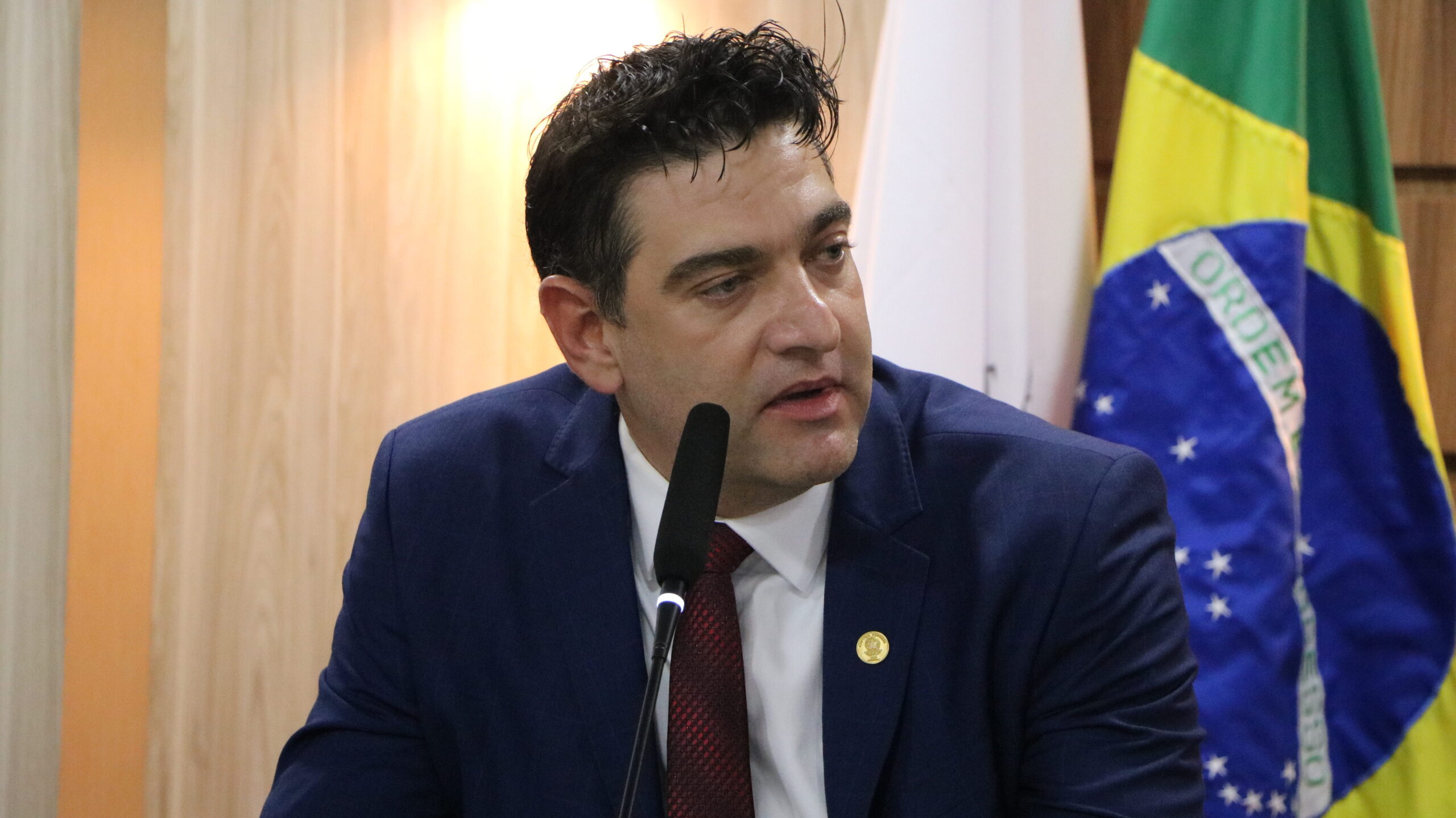 Um close-up de um homem, vestido com traje formal (terno e gravata), falando em um microfone de mesa. Ele está em um ambiente interno com painéis de madeira clara no fundo. À direita, é possível ver duas bandeiras hasteadas.