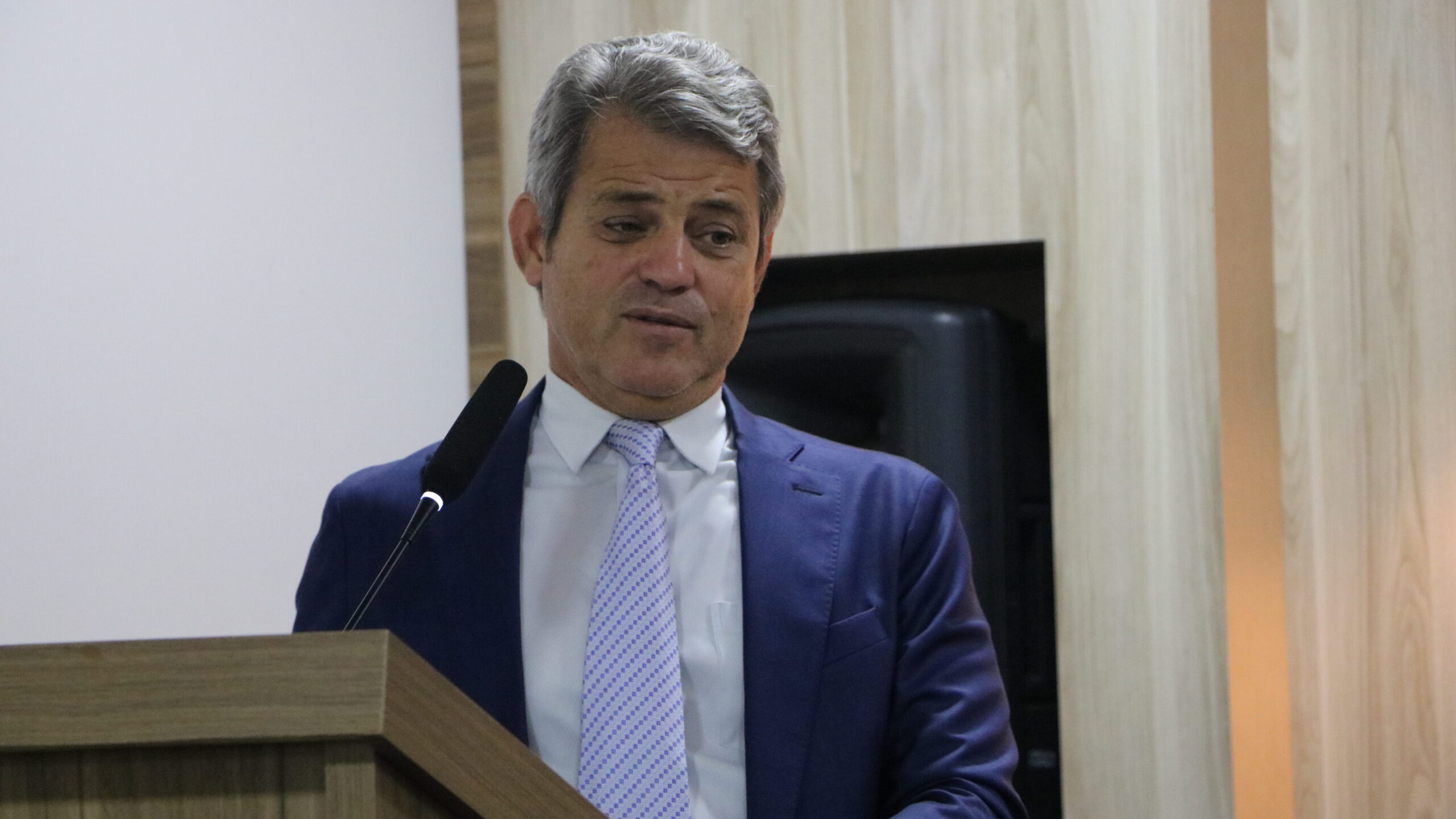 Um close-up de um homem maduro, vestido com traje formal (terno e gravata), em um púlpito de madeira. Ele está em um ambiente interno.