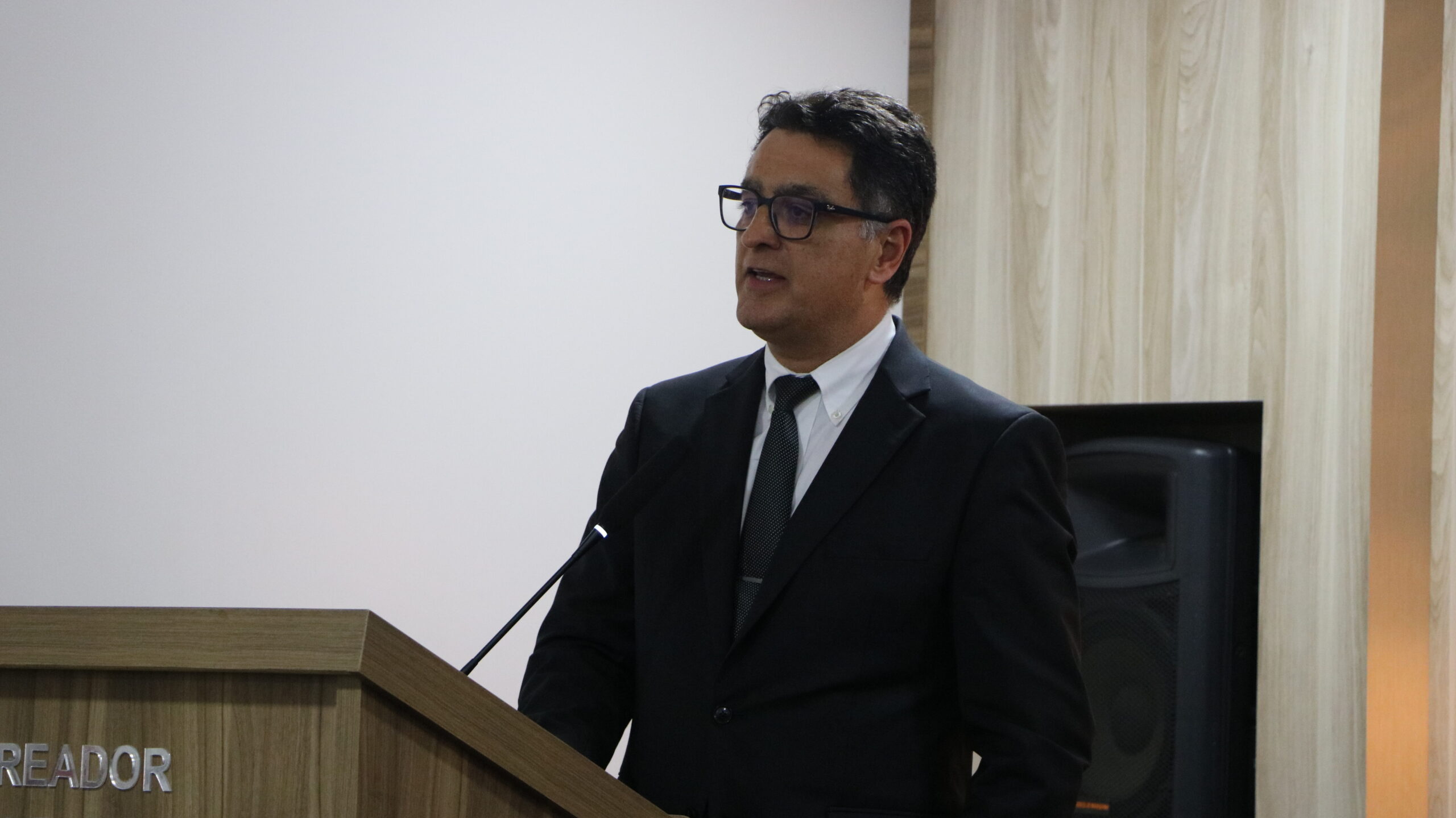 Um close-up de um homem de óculos, vestido com traje formal, enquanto fala em um púlpito de madeira. Ele está posicionado em frente a uma parede branca lisa. Um alto-falante escuro está visível no painel de madeira à direita.