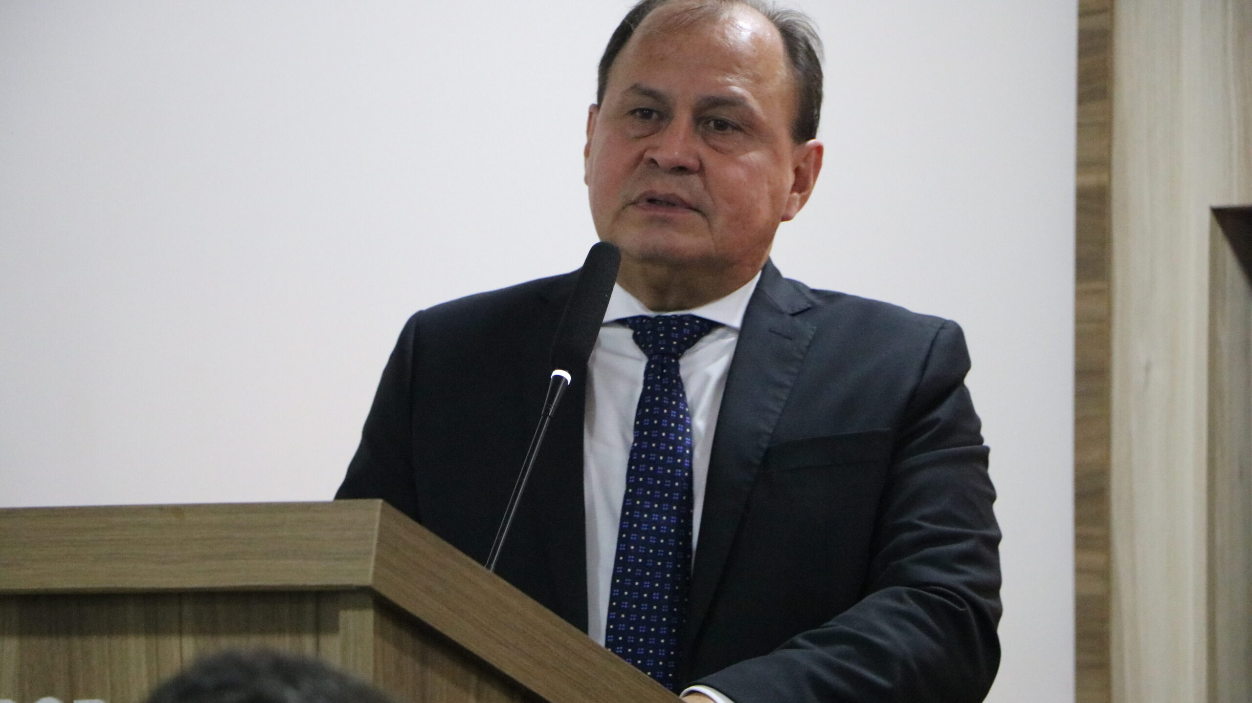 Um close-up de um homem, vestido com traje formal (terno e gravata), enquanto fala ao microfone. Ele está em um púlpito de madeira. O fundo é simples, consistindo em uma parede branca e um revestimento de madeira na lateral, destacando o orador.