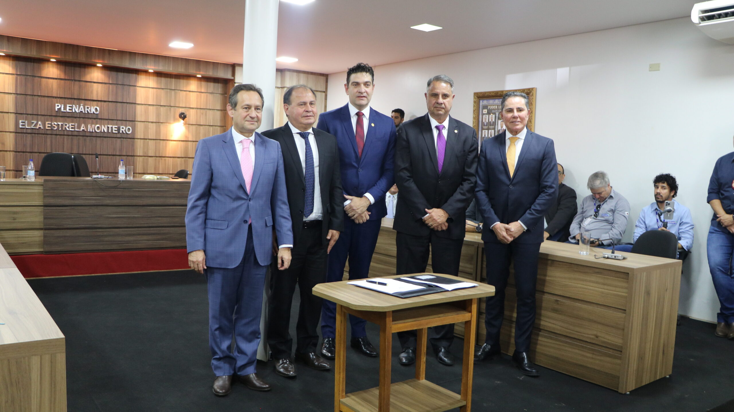 Cinco homens, todos vestidos com trajes formais, estão posando em pé em um ambiente interno. No centro, há uma pequena mesa de madeira clara com alguns documentos. O fundo é dominado por uma grande mesa de madeira escura e uma parede de madeira onde se lê "PLENÁRIO ELZA ESTRELA MONTEIRO".