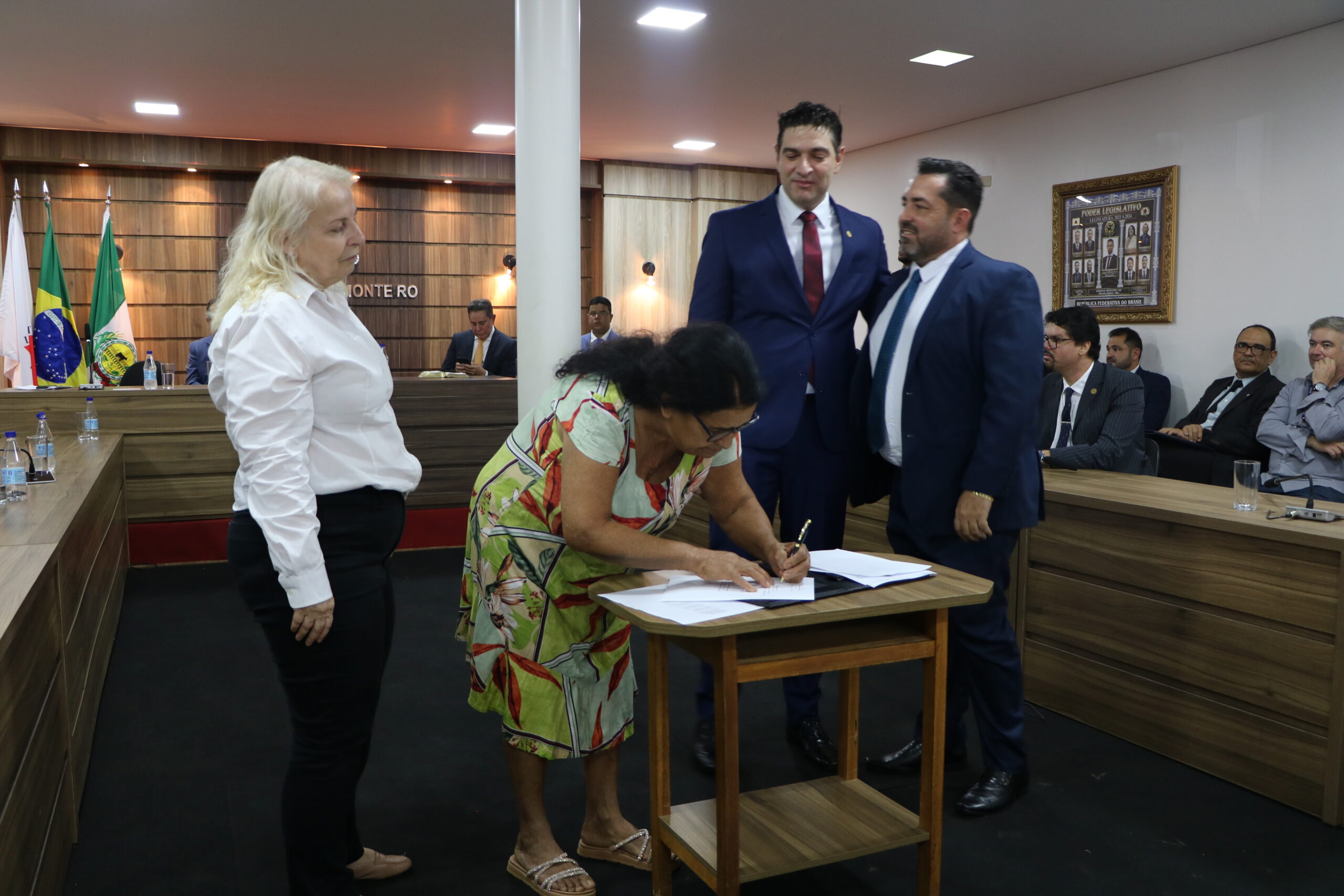 Uma cena de uma cerimônia em um ambiente formal. Uma mulher, vestida de forma social e colorida, está assinando documentos em uma pequena mesa de madeira, enquanto outras duas pessoas, vestidas de forma formal, a observam. O fundo da sala apresenta bandeiras e uma placa com o nome de uma sala.