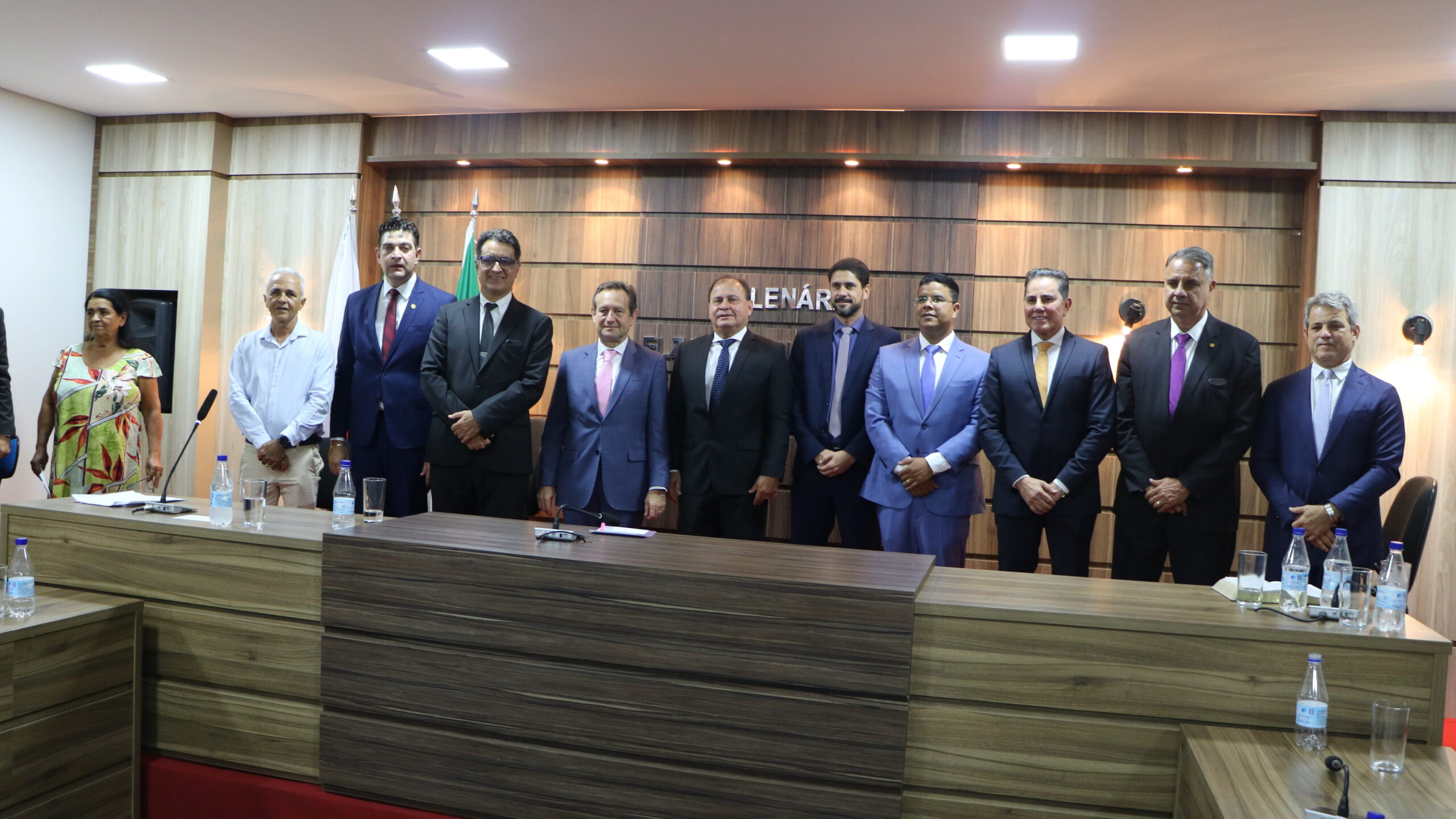 Uma foto de grupo tirada em um ambiente interno formal. Um grupo de dez pessoas está posando em pé atrás de uma mesa grande de madeira escura. A maioria dos homens veste trajes formais, e há uma mulher à esquerda em trajes sociais. A parede de fundo é revestida de madeira.