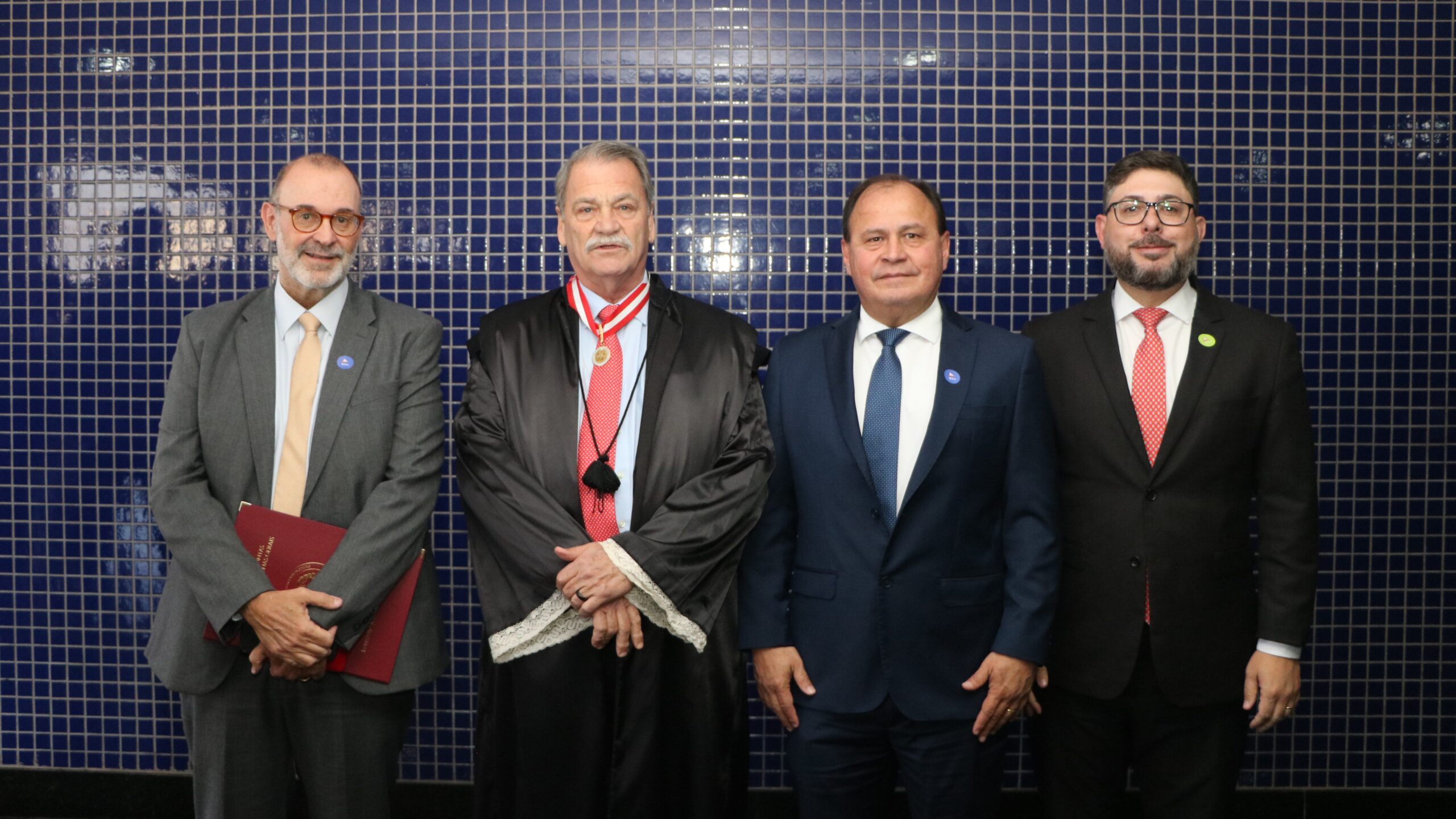 Grupo de quatro homens posando lado a lado em um evento formal, todos usando medalhas no pescoço. O segundo homem veste beca sobre seu traje, e o fundo é uma parede revestida de pequenos azulejos.