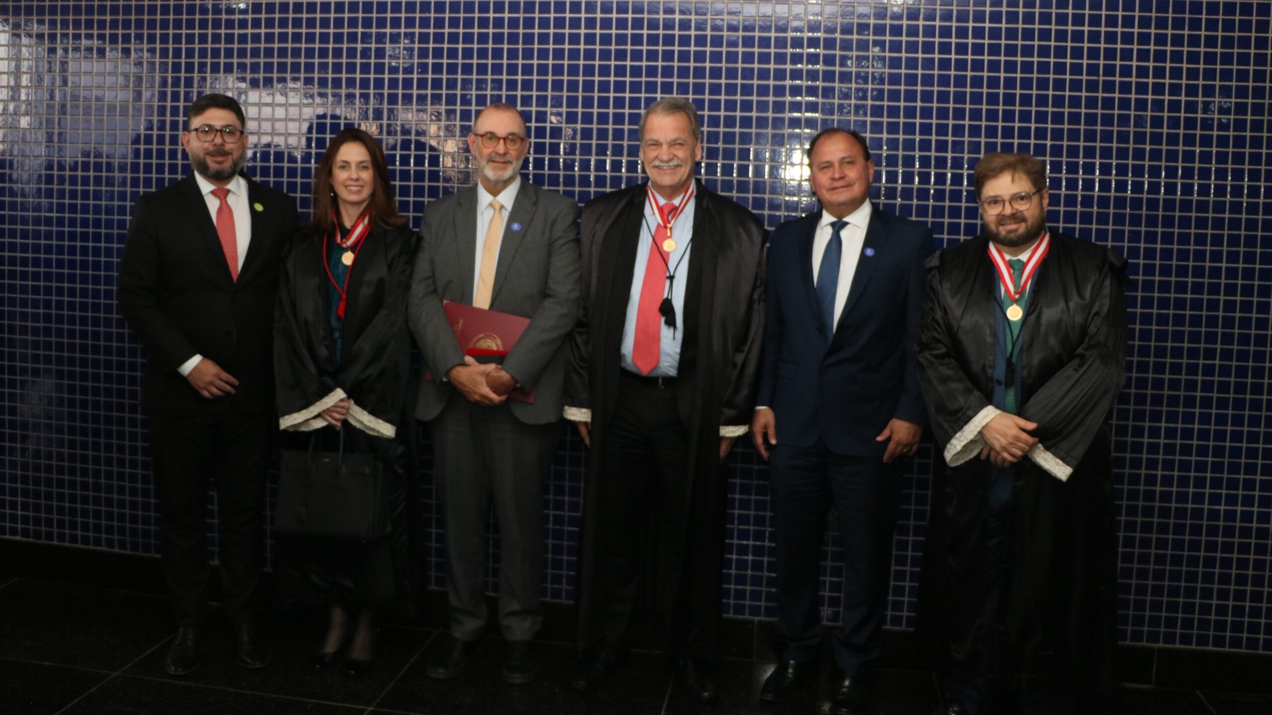 Grupo de seis pessoas (cinco homens e uma mulher) posando lado a lado em um evento formal, todos usando medalhas no pescoço. Três homens e a mulher vestem becas sobre seus trajes, e o fundo é uma parede revestida de pequenos azulejos.