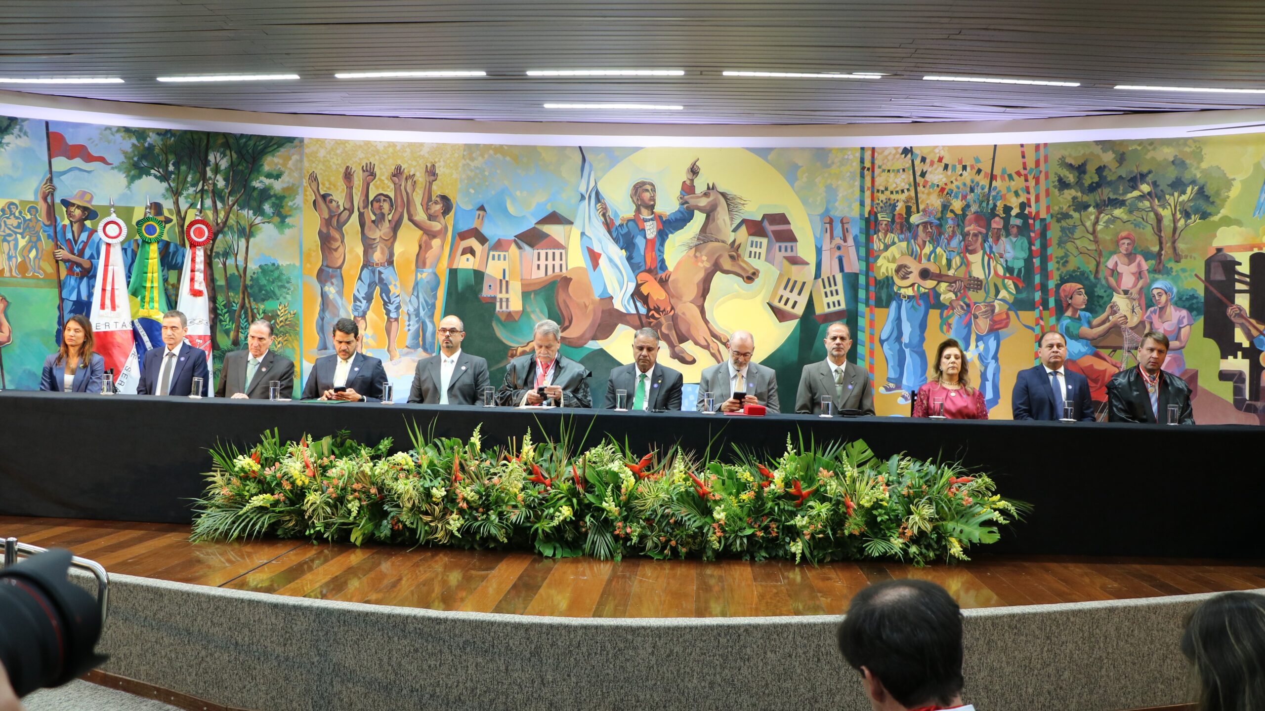 Mesa de honra em um auditório, com 12 pessoas sentadas em vestimentas formais e sociais, posicionadas atrás de uma longa mesa decorada com um arranjo de flores e folhagens. Ao fundo, um grande painel artístico retrata cenas culturais e históricas, com bandeiras nas laterais.