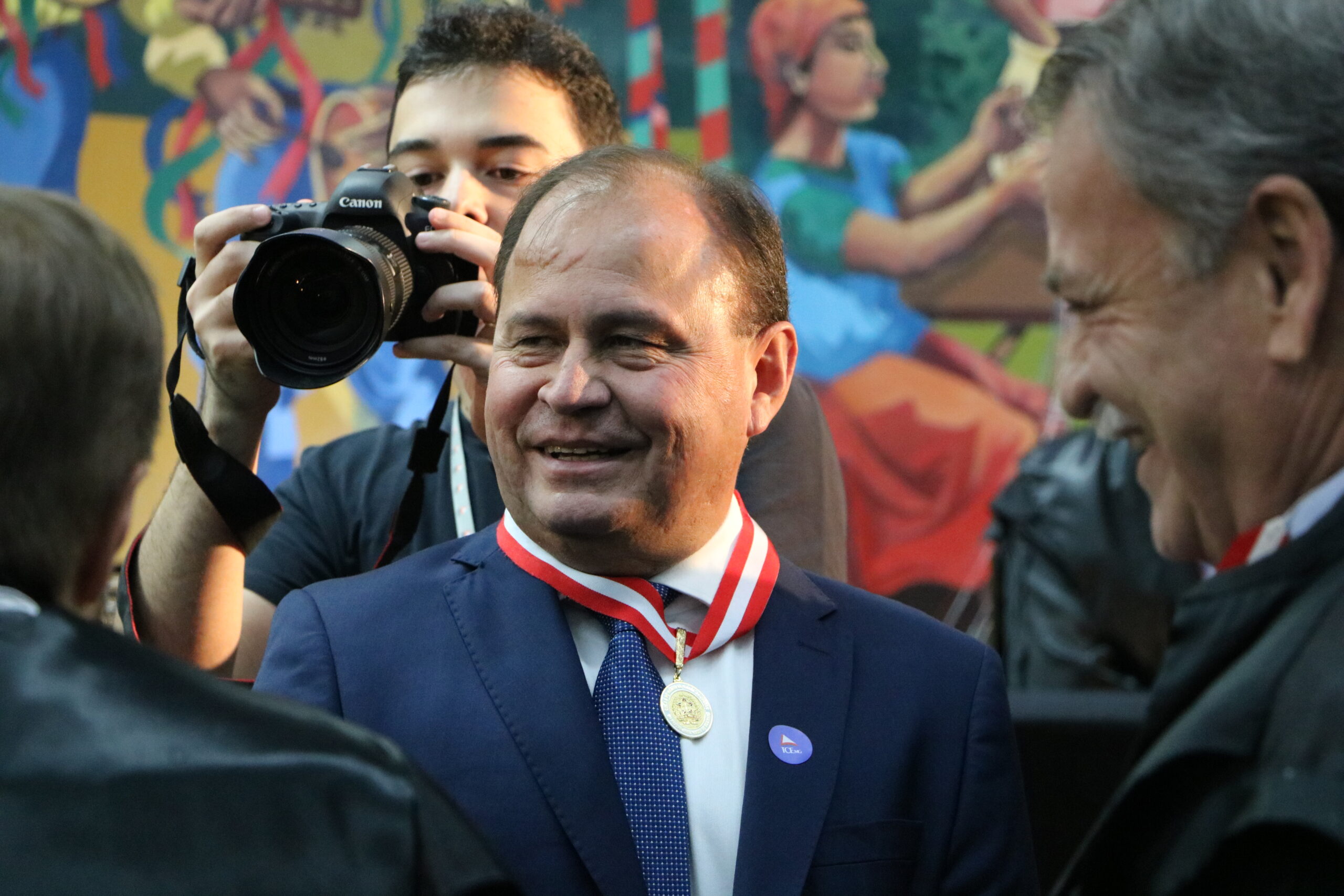 Retrato de um homem de vestes formais e medalha no pescoço, sorrindo e sendo fotografado em um momento informal do evento.