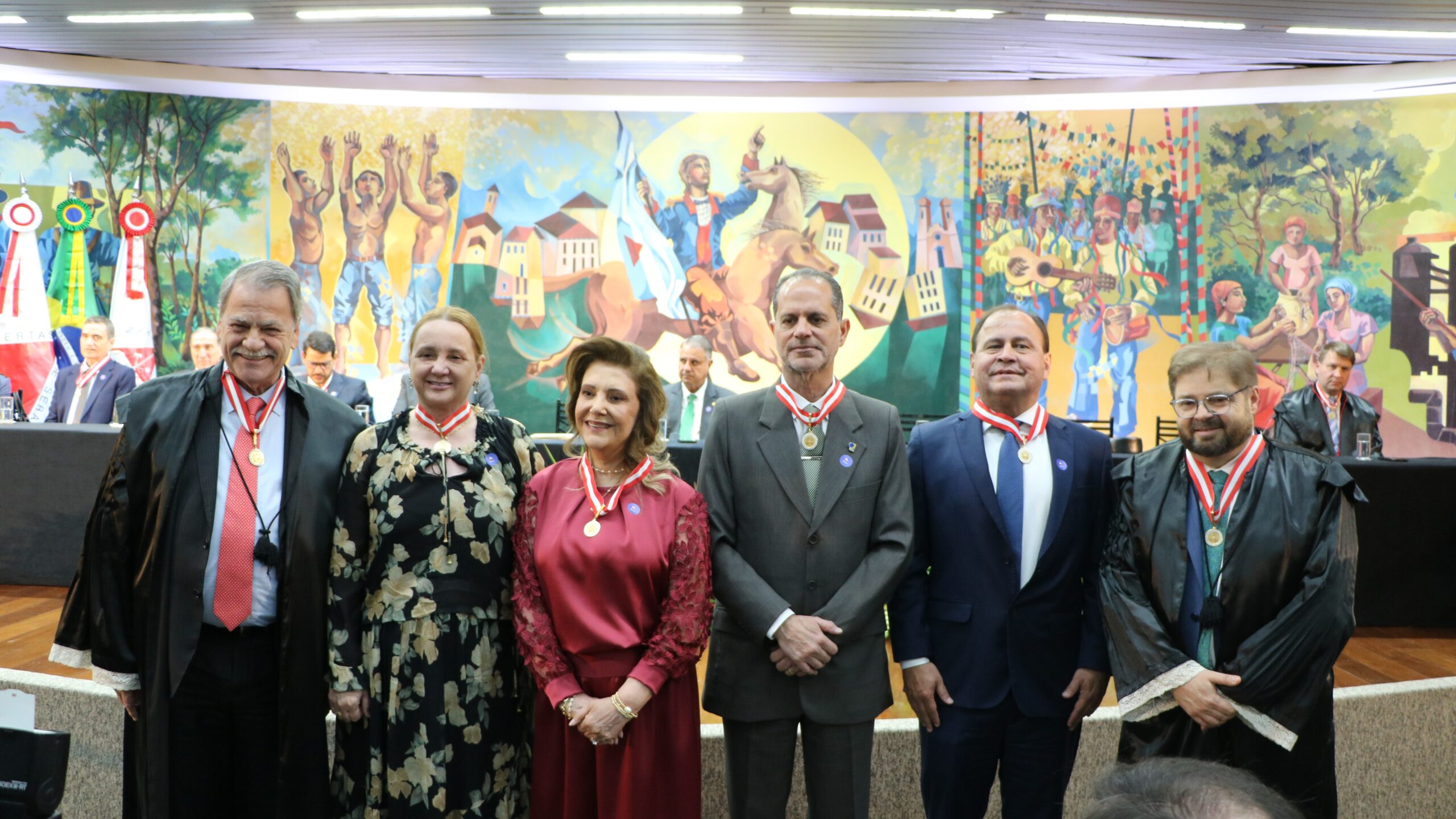 Foto de grupo de seis pessoas (quatro homens e duas mulheres) em um evento de homenagem ou premiação, todas usando medalhas penduradas em fitas no pescoço. O grupo está posicionado à frente de um grande painel artístico.