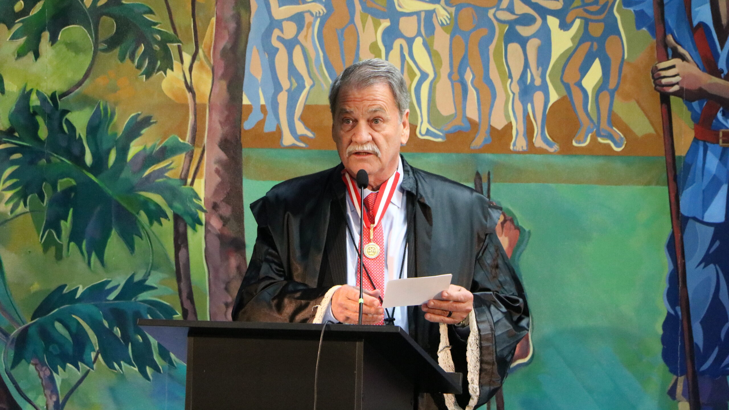 Retrato de um homem em discurso, usando beca e uma medalha no pescoço, posicionado atrás de um púlpito escuro. Ele segura um pedaço de papel na mão. O fundo é um grande painel artístico com figuras humanas e folhagens.