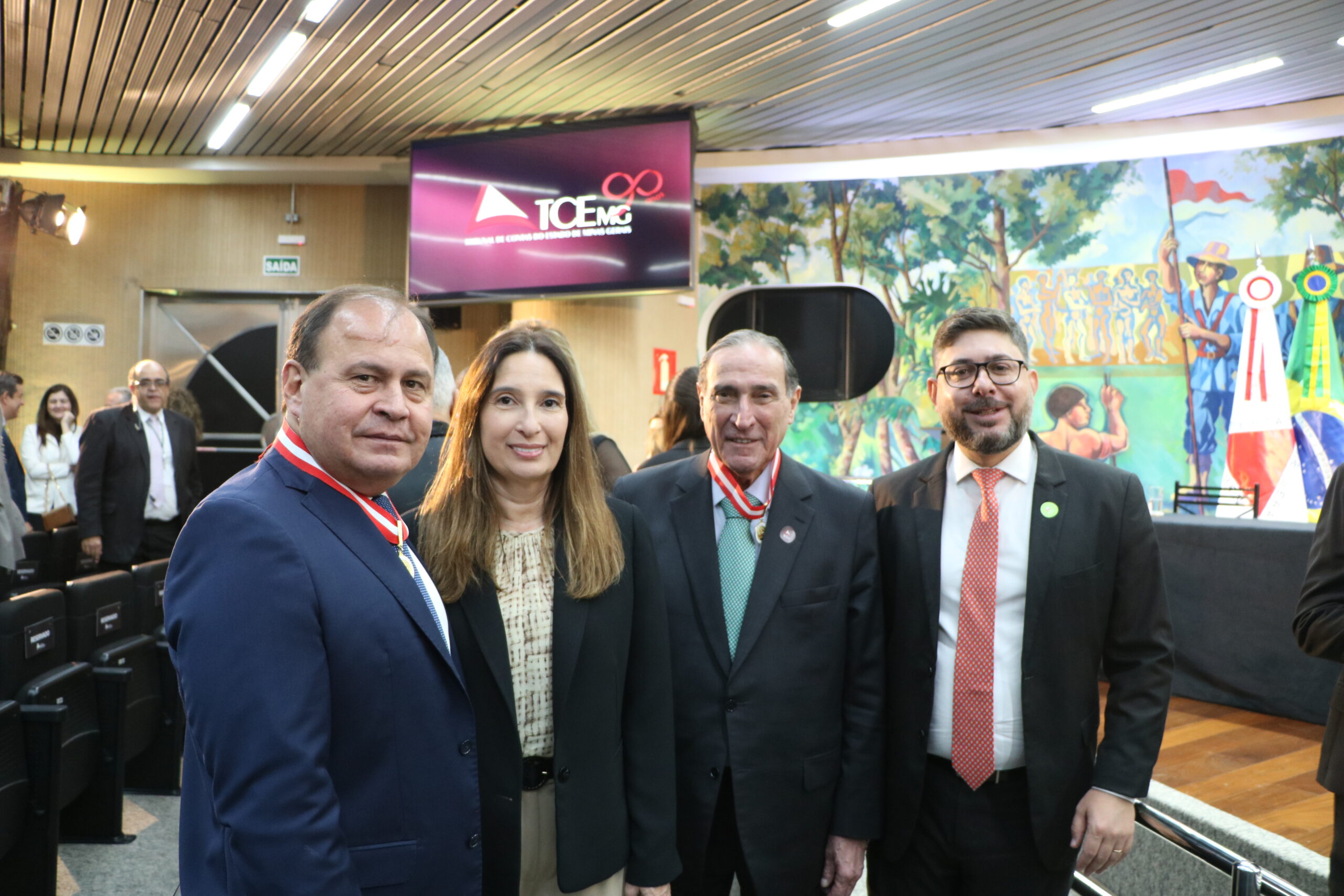 Grupo de quatro pessoas (três homens e uma mulher) posando em ambiente interno, contra um fundo que inclui um painel artístico, uma tela de apresentação e outras pessoas ao fundo.