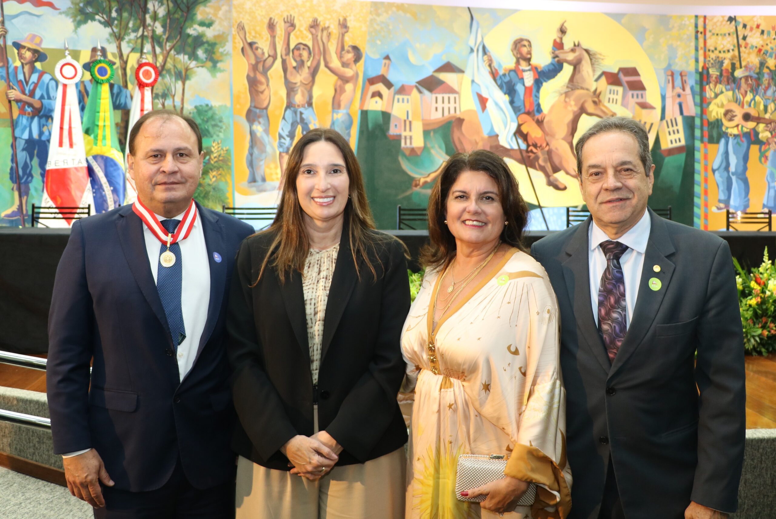 Grupo de quatro pessoas (dois homens e duas mulheres) posando em pé à frente de um painel artístico com temática histórica e cultural. Os homens usam trajes formais e as mulheres vestem trajes sociais e executivos.