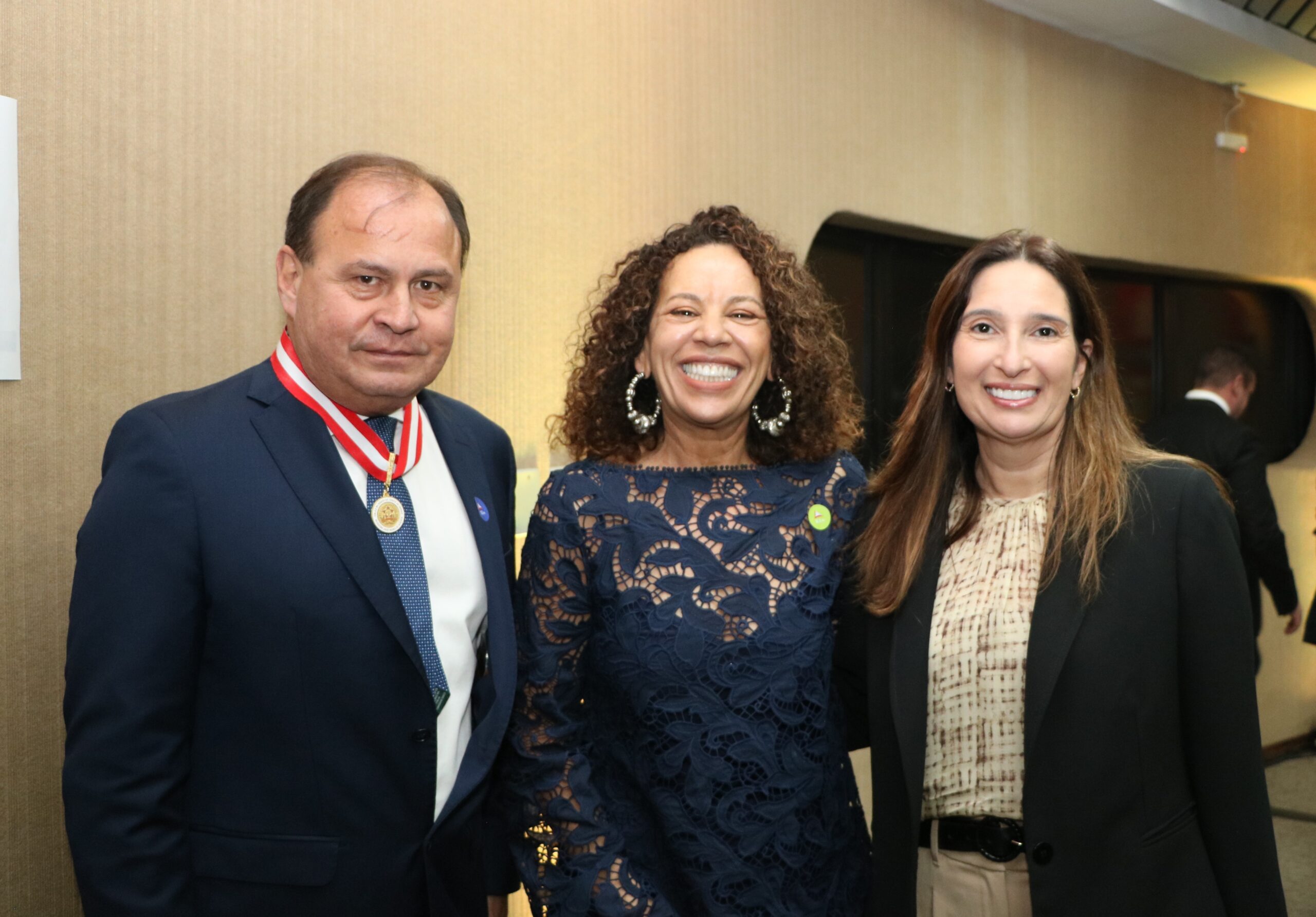 Foto de um homem e duas mulheres posando lado a lado em evento formal. O homem veste traje formal e medalha no pescoço, e as mulheres usam trajes sociais/executivos.