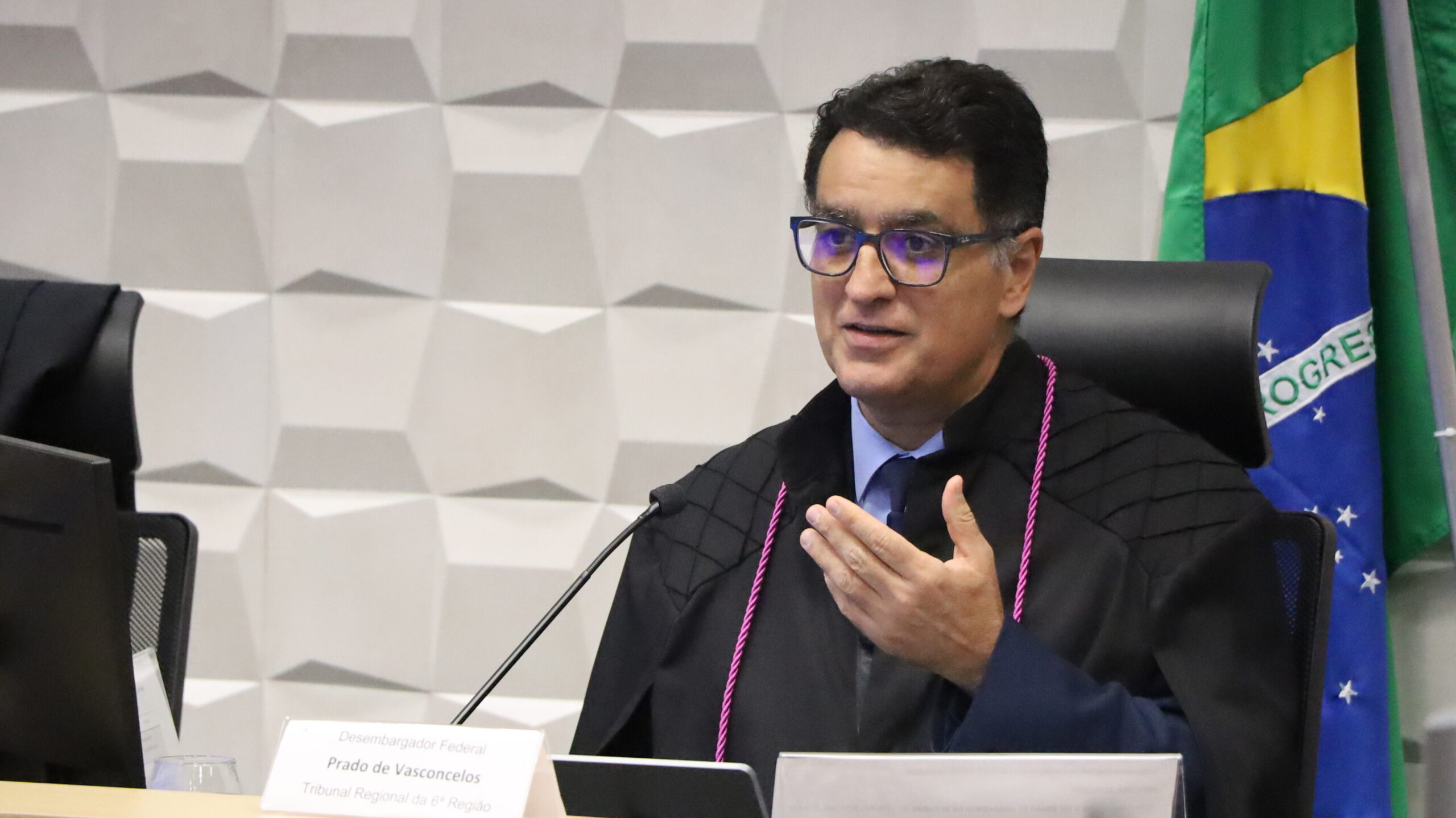 Desembargador federal Prado de Vasconcelos, de óculos e vestindo toga, sentado em uma mesa de tribunal, gesticula enquanto fala, com a bandeira do Brasil ao fundo.