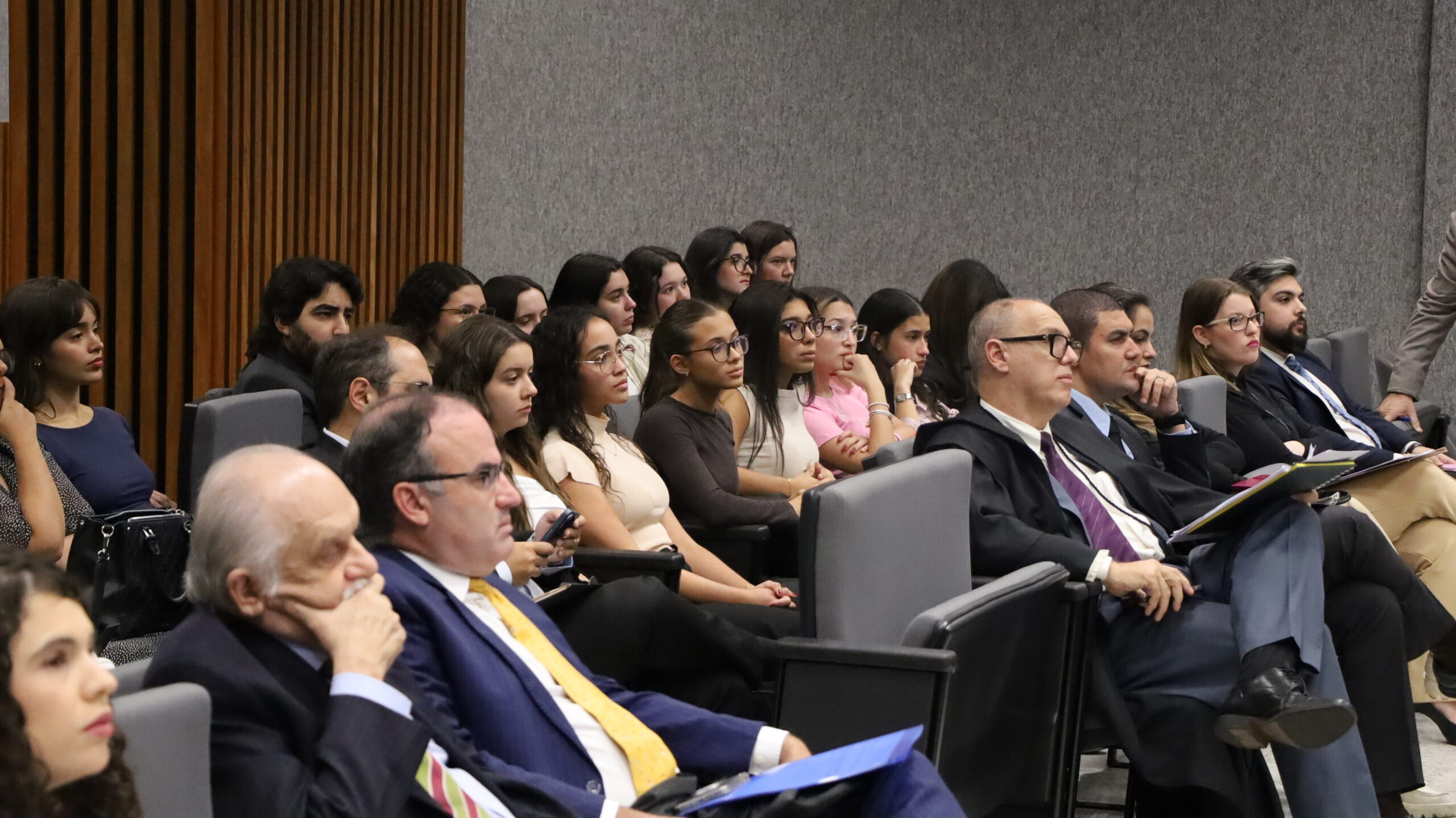 Pessoas em um auditório, incluindo jovens e adultos, em trajes formais e casuais, sentadas em cadeiras acolchoadas, atentas a uma apresentação ou discurso.