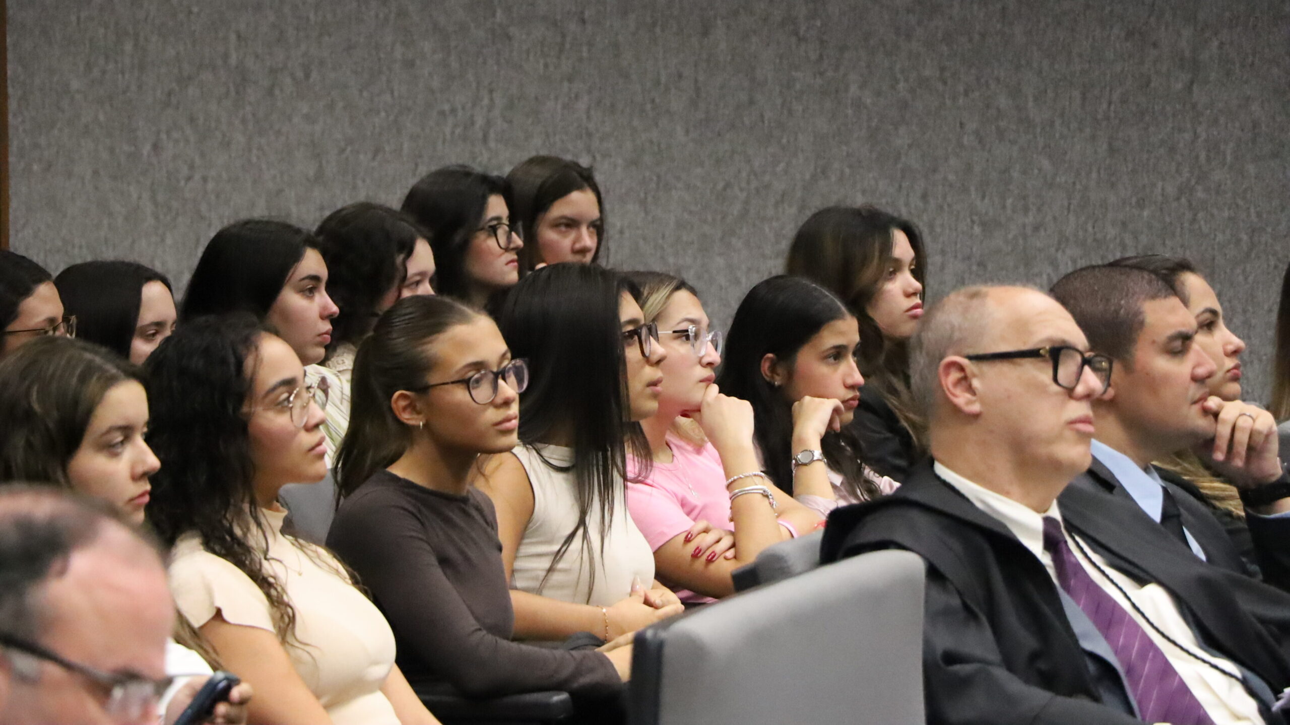 Pessoas, principalmente jovens mulheres, sentadas em um auditório, prestando atenção a um evento ou apresentação.