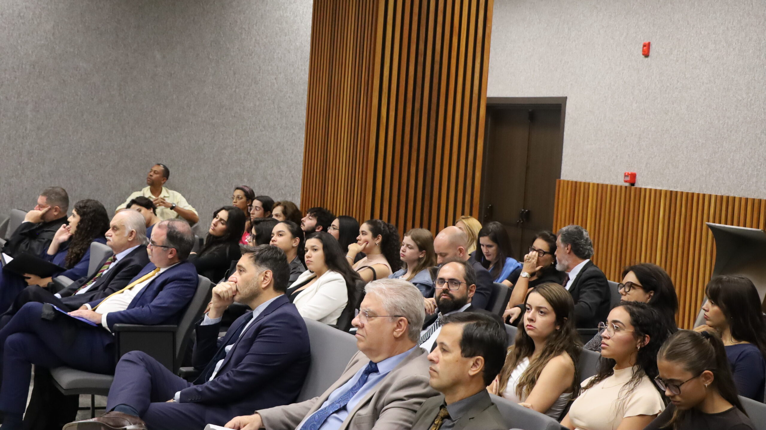 Audiência atenta sentada em cadeiras de auditório, com painéis de madeira e parede cinza ao fundo.