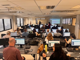 A imagem mostra várias fileiras de mesas com computadores se estendem pela sala. Os participantes estão sentados, olhando para os monitores ou para a frente. No fundo, é possível ver uma mulher de pé, interagindo com o grupo próximo a uma tela de projeção ou quadro. As salas possuem iluminação interna e paredes claras.