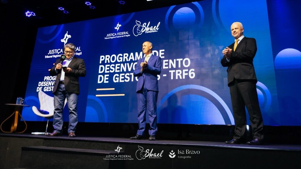 Três homens, todos vestidos de forma formal, estão em um palco elevado. O palco é iluminado com luzes de destaque e tem uma tela grande e brilhante ao fundo. A tela exibe o texto "PROGRAMA DE DESENVOLVIMENTO DE GESTORES - TRF6" e os logotipos de instituições. O homem à esquerda está segurando um microfone, o homem ao centro está com as mãos juntas, e o homem à direita está aplaudindo.