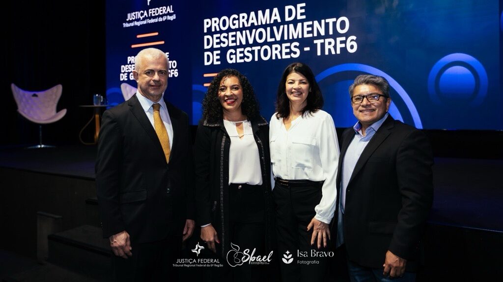 Quatro pessoas, incluindo um homem vestido de forma formal e três mulheres com trajes sociais, posam juntas em um palco com uma grande tela iluminada ao fundo. A tela exibe o texto "PROGRAMA DE DESENVOLVIMENTO DE GESTORES - TRF6" e logotipos institucionais.
