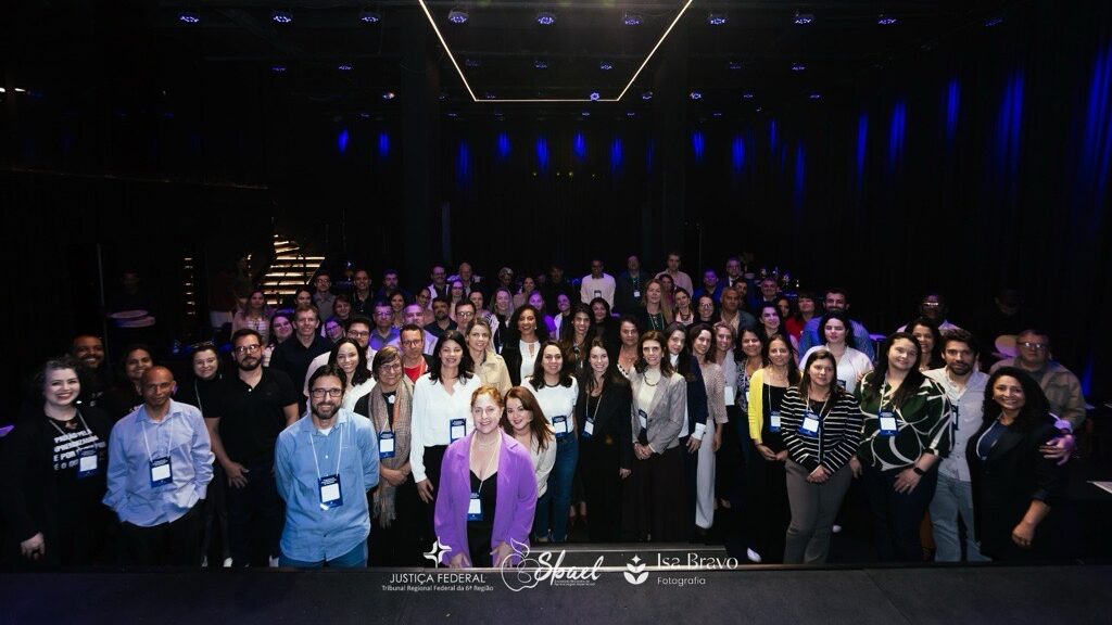 Esta é uma foto em grupo tirada em um ambiente interno com iluminação baixa, com iluminação de destaque azul no fundo. Um grande número de pessoas está posando, alinhadas em várias filas, olhando para a câmera. Estão vestidas de forma casual e semi-formal, e a maioria usa crachás. Há textos sobrepostos na parte inferior que incluem o nome de uma instituição e de uma fotógrafa.