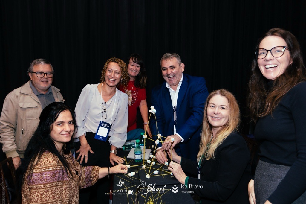 Um grupo de cerca de sete pessoas, vestidas em trajes casuais e semi-formais, sorri e interage em volta de uma mesa.