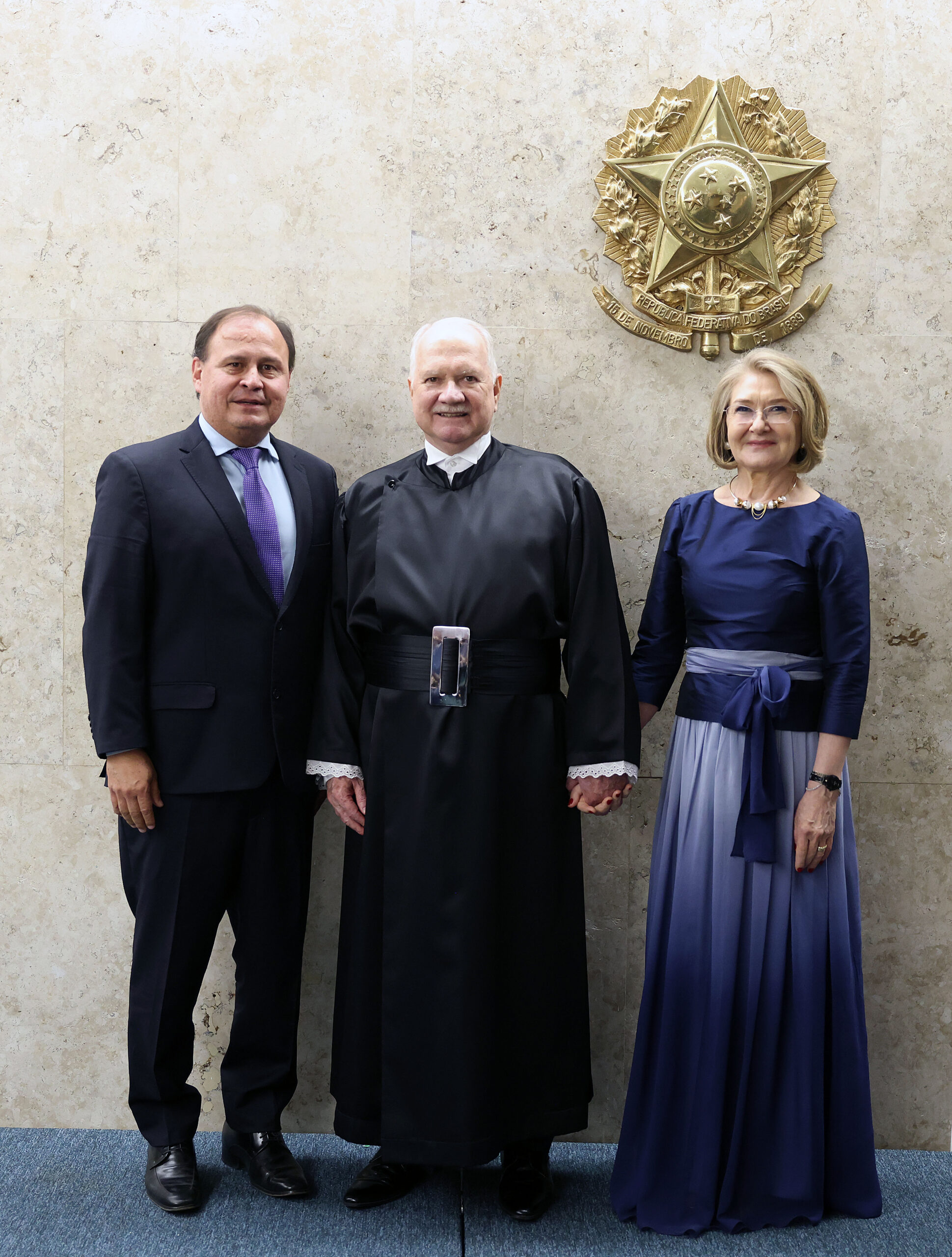 Três pessoas posando para uma foto em pé. No centro, um homem de toga preta está ladeado por um homem de terno escuro e uma mulher de vestido azul, em frente a uma parede de mármore com o Brasão da República em relevo dourado.