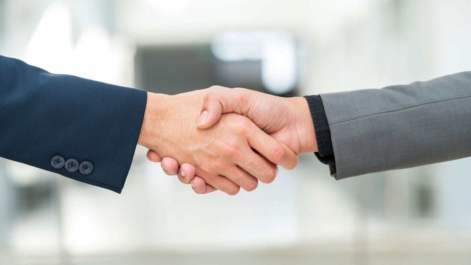 A imagem é um close de um aperto de mãos entre duas pessoas. A pessoa à esquerda usa um terno azul-marinho, e a pessoa à direita, um terno cinza. O foco está no aperto de mão, que parece ser firme. O fundo é desfocado e parece ser um corredor ou saguão bem iluminado de um escritório. A imagem transmite a ideia de um acordo, parceria ou cumprimento profissional.