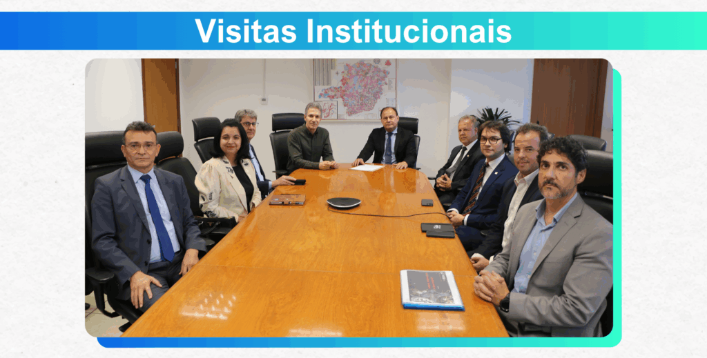 A imagem é um slide de apresentação com o título "Visitas Institucionais" em um fundo azul. A foto principal mostra um grupo de nove pessoas sentadas ao redor de uma grande mesa de reunião de madeira. Eles estão vestindo trajes formais.

Todos olham para a câmera, e alguns sorriem. Na parede atrás deles, há um mapa e uma televisão. A iluminação é clara e o ambiente, de escritório. Sobre a mesa, há papéis, um microfone e um livro.